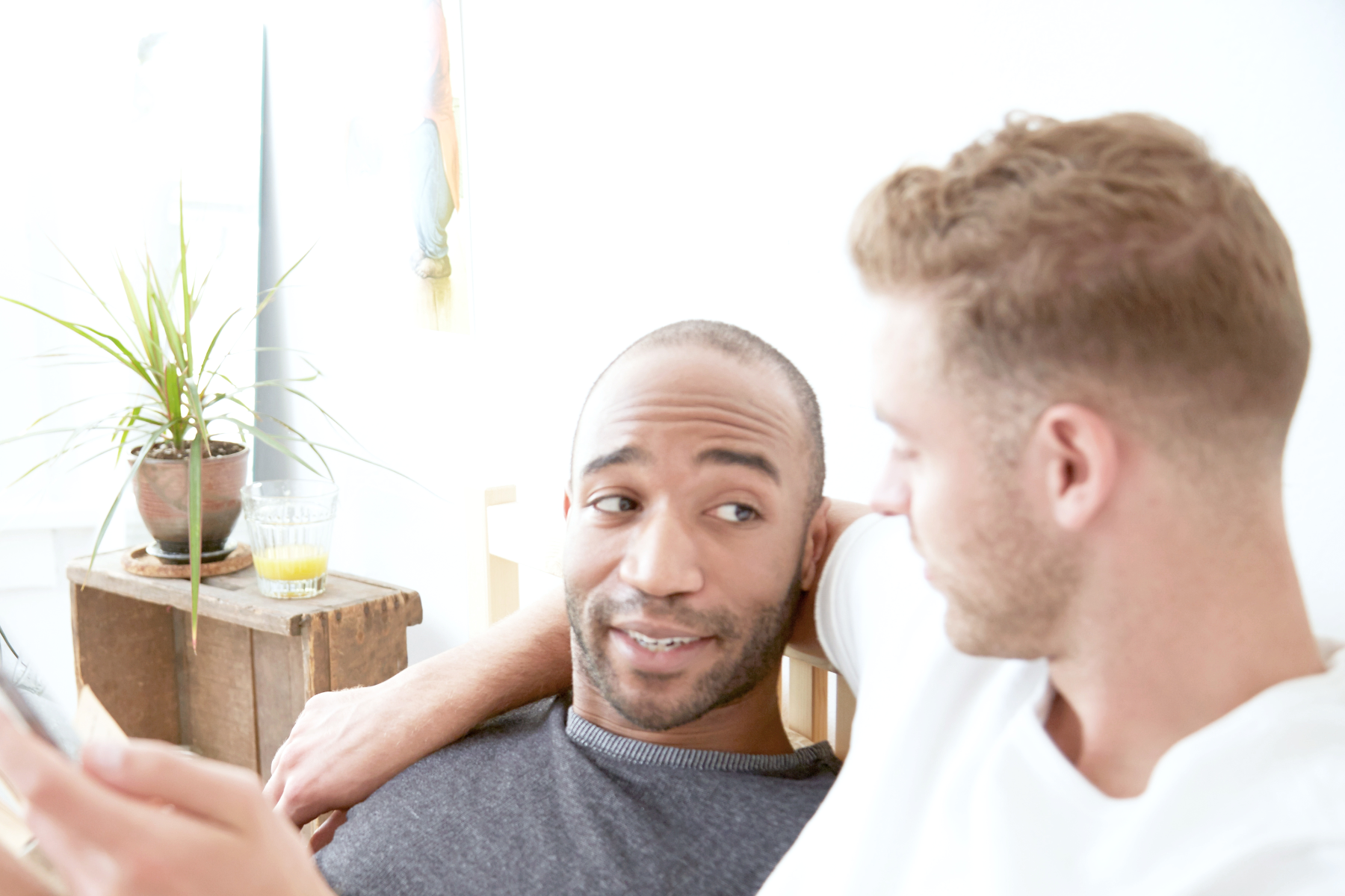 Two men sitting on a bed, one with his arm around the other, smiling and looking at each other