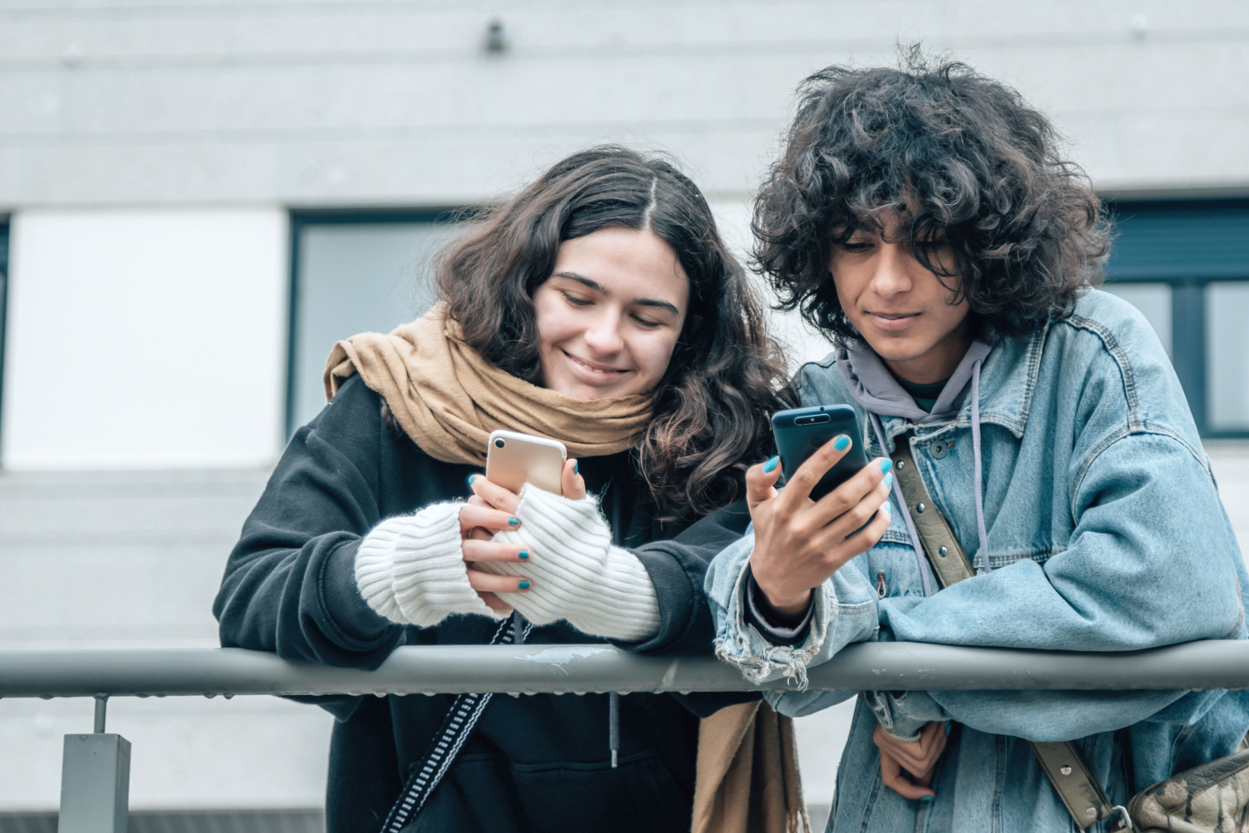 Two teenagers, a girl and a boy, are standing outside, leaning on a railing, looking at their phones and smiling.