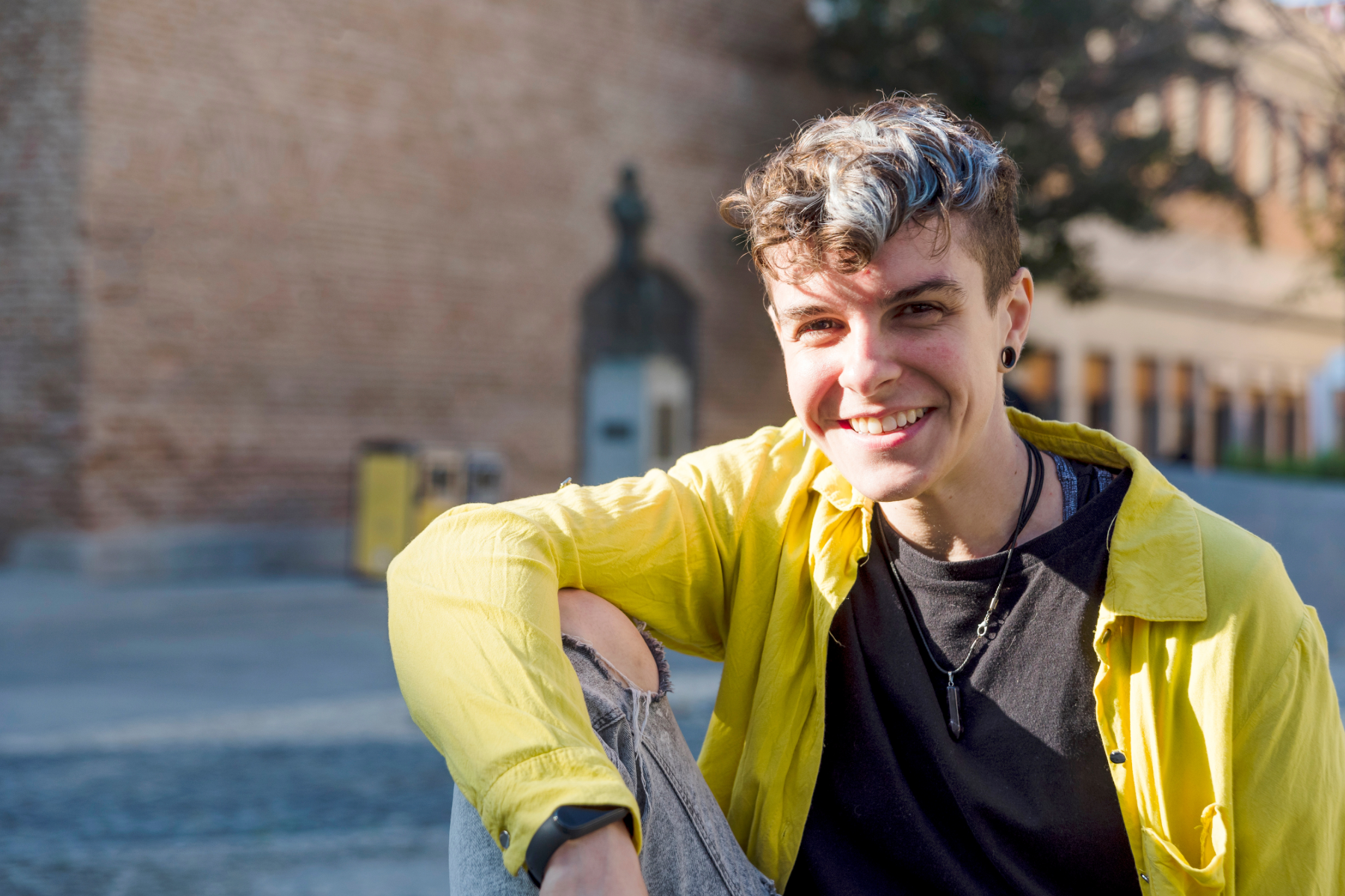 Young person with short curly hair, smiling, sitting outdoors in front of a brick wall, wearing a yellow jacket, black T-shirt, and accessories.