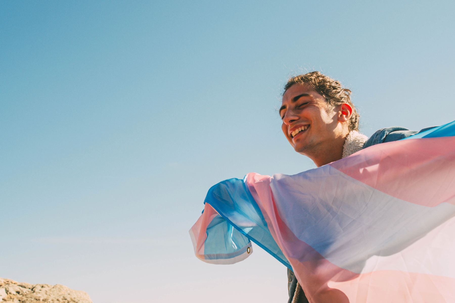 A young man smiling and laughing outdoors on a clear, sunny day with a bright blue sky, holding a colorful, semi-transparent fabric or flag.