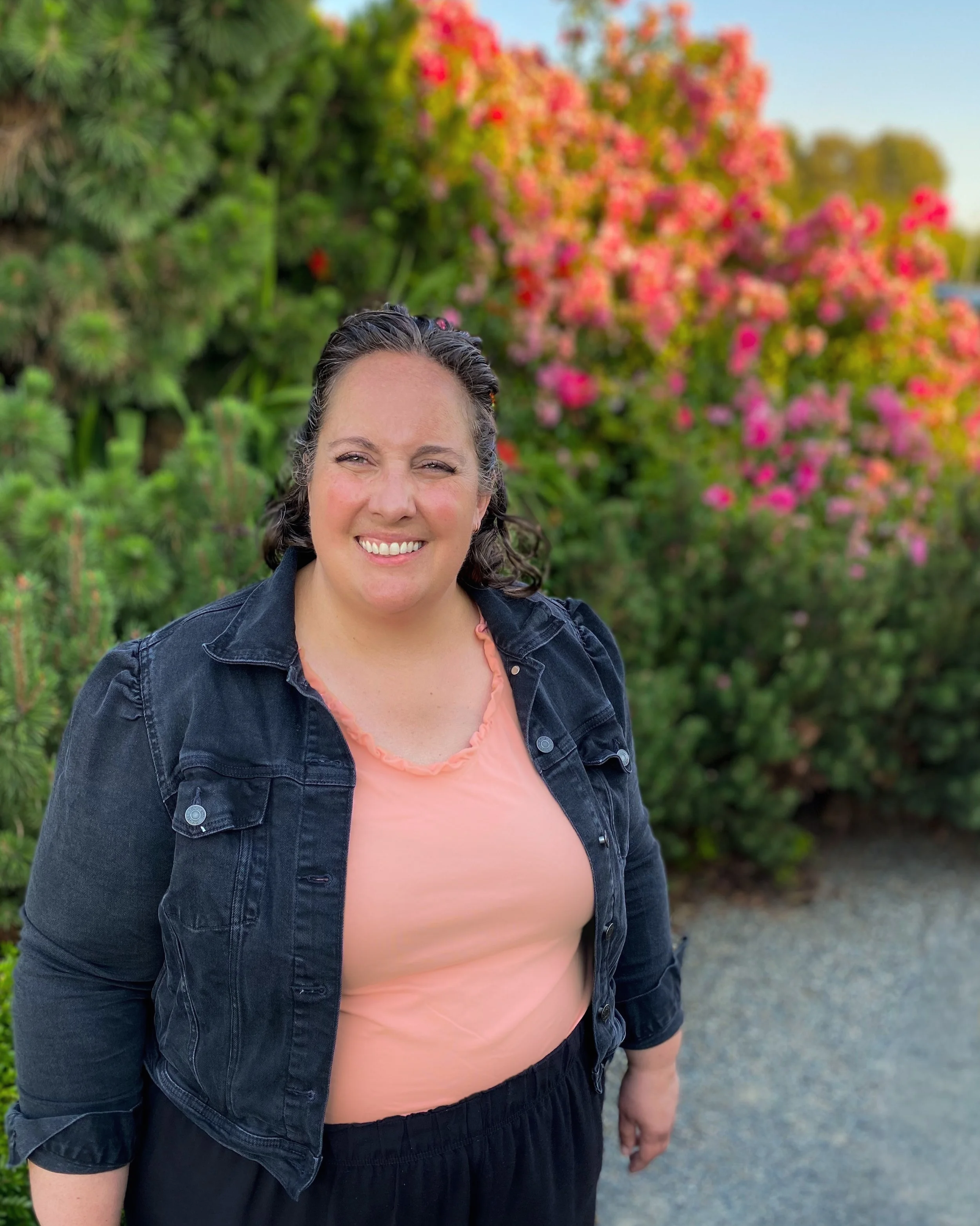A woman smiling outdoors with pink and orange flowering bushes in the background.