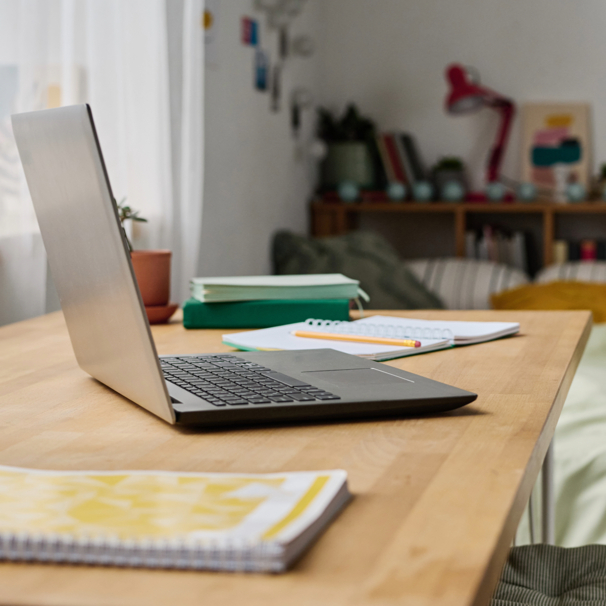 A laptop on a wooden desk with notebooks, a pencil, and a plant, in a cozy room with shelves and decorations in the background.