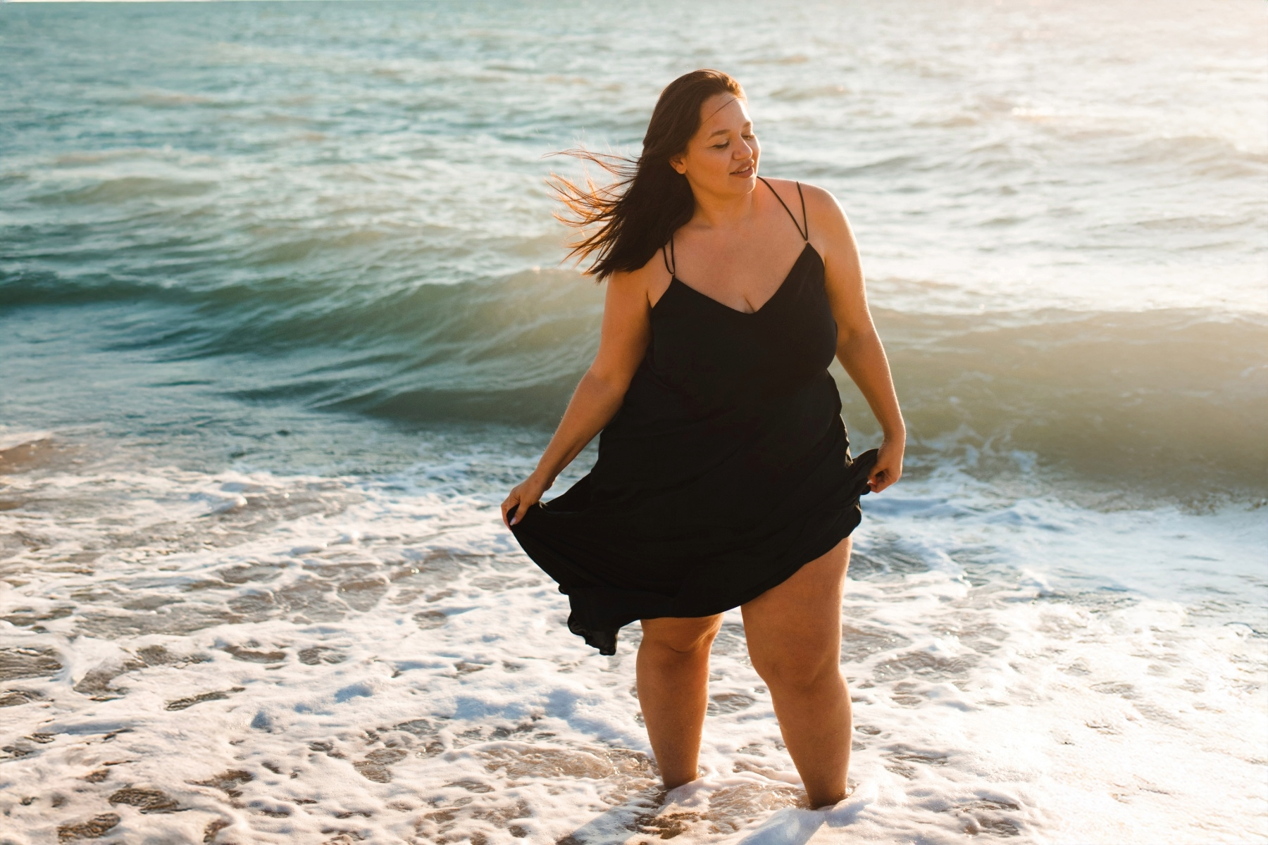 Woman in a black dress standing in the ocean with waves crashing around her during sunset.