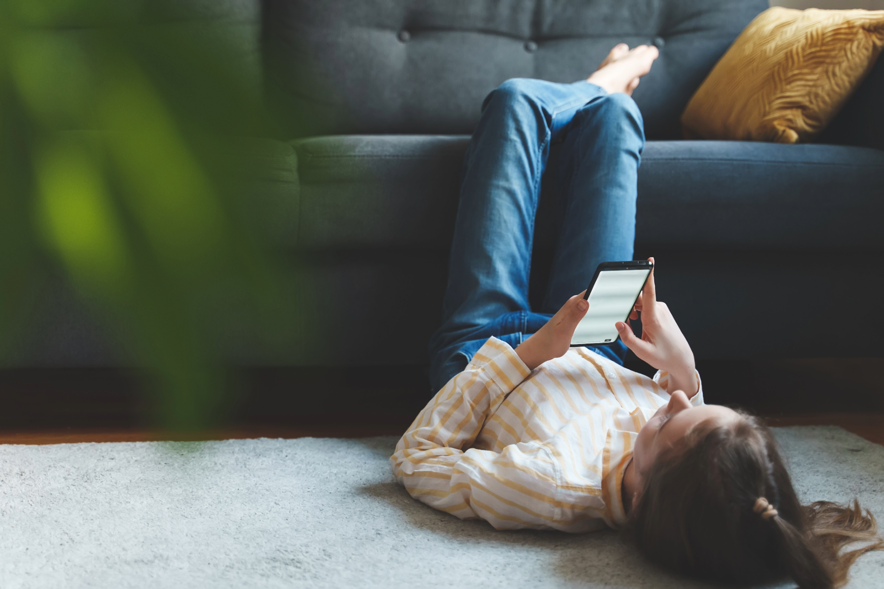 Woman lying on her back on a carpeted floor, looking at her smartphone, with legs extended on a sofa in the background, and a yellow pillow on the sofa.