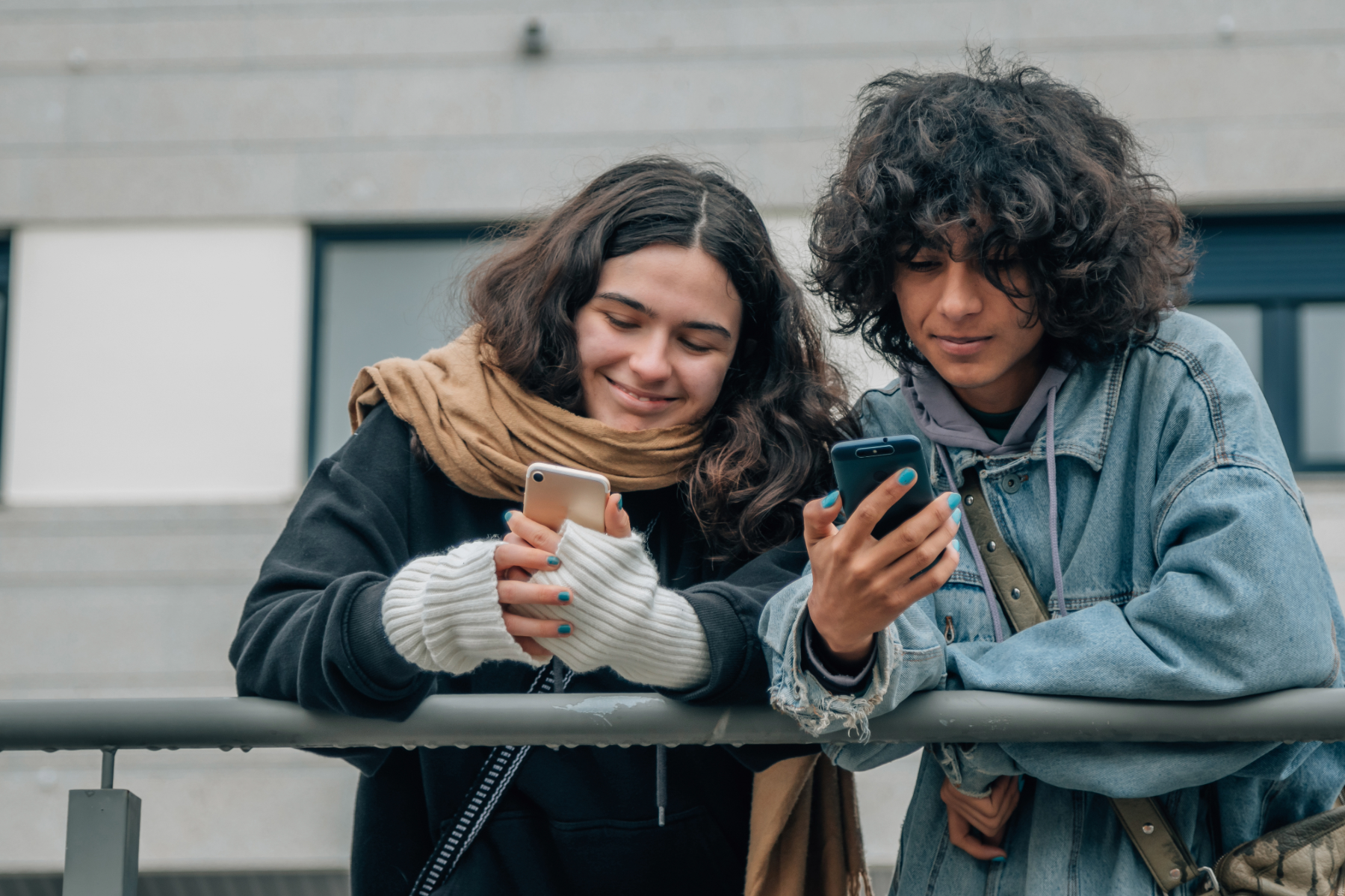 Two teenagers, a girl and a boy, are standing outside, leaning on a railing, looking at their phones and smiling.
