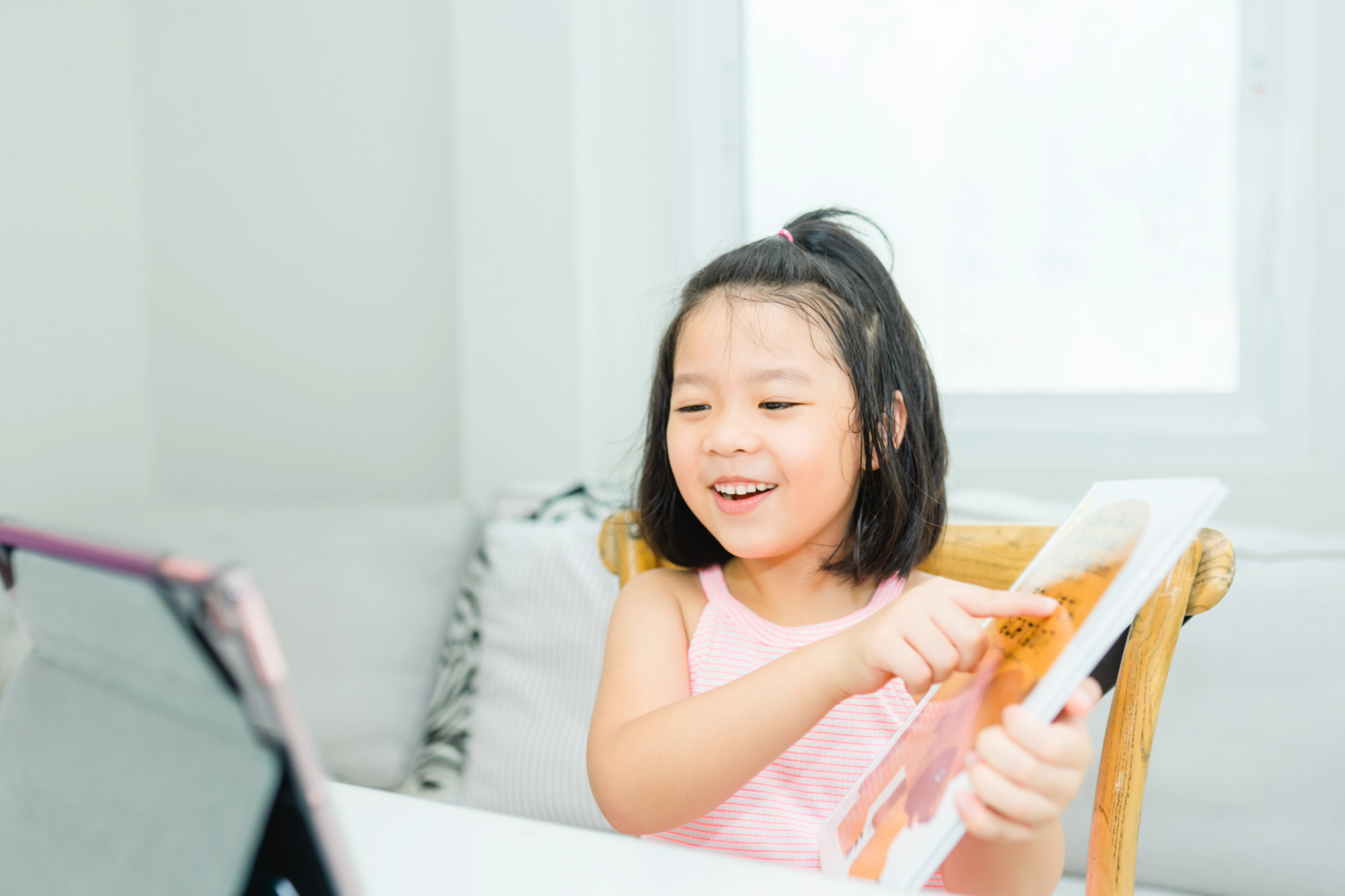 A young girl with black hair tied up with a pink hair tie, wearing a pink and white striped sleeveless top, smiling while reading a book. She is sitting on a wooden chair near a white table, with a tablet in front of her. The background features a window and light-colored walls.
