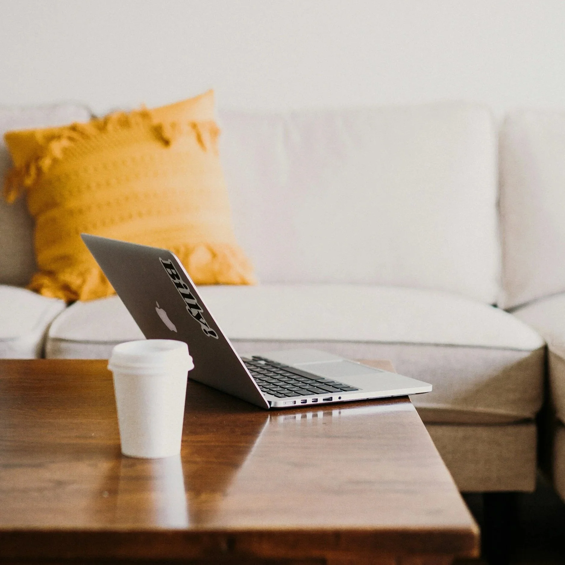 A laptop on a wooden table with a white coffee cup in front of it, and a white sofa with a yellow pillow in the background.