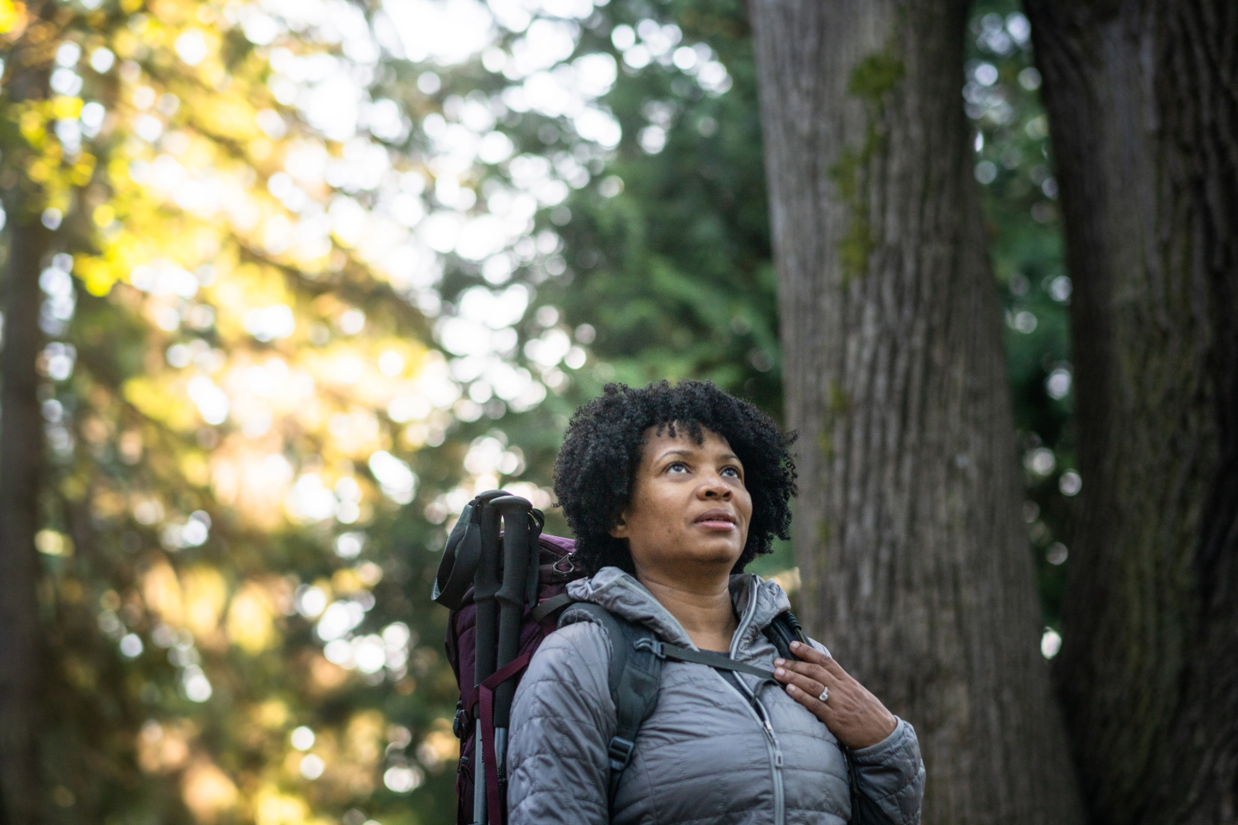 Woman with a backpack in a forest, looking upwards with a thoughtful expression.