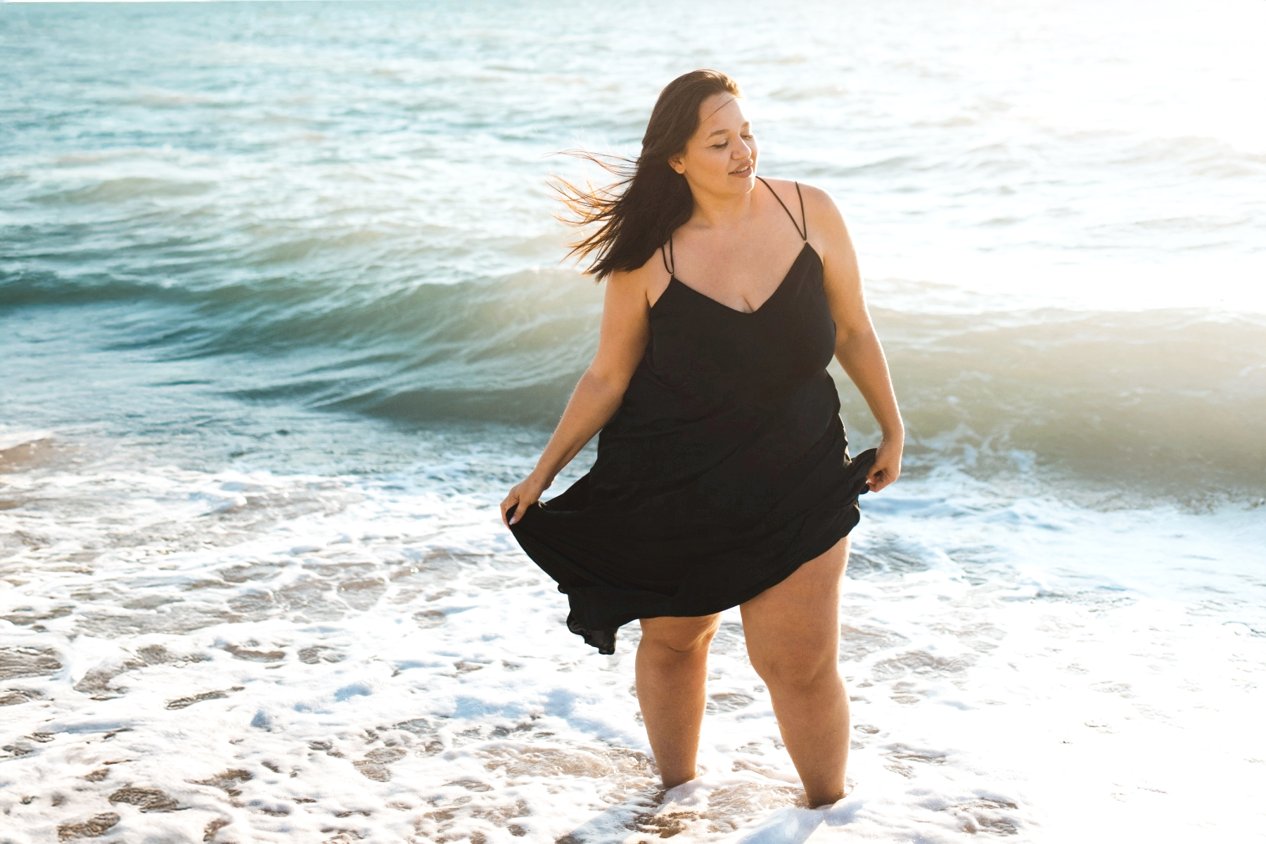 Woman in a black dress standing in the ocean with waves crashing around her during sunset.