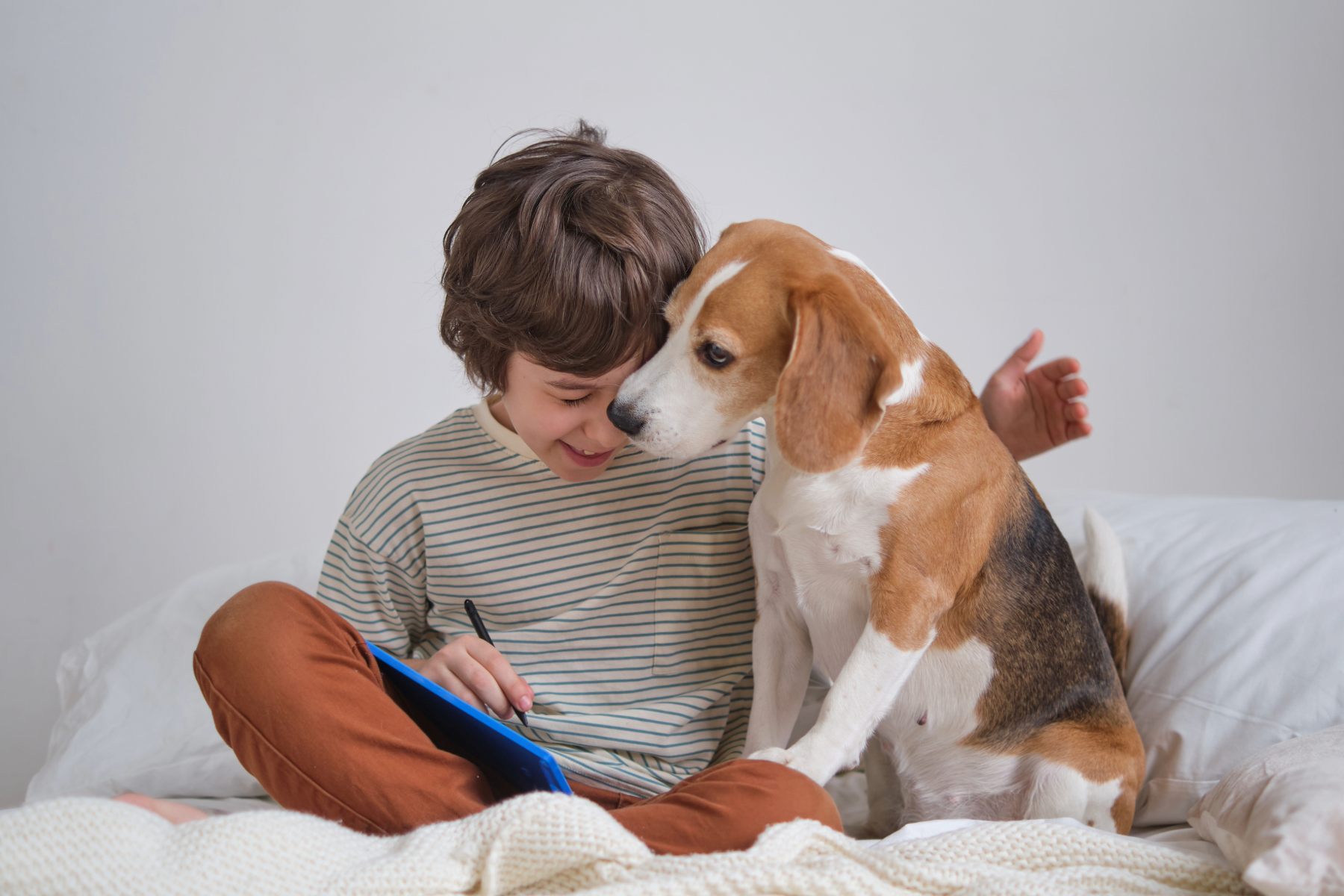 A young boy with brown hair, smiling, sitting on a bed with a beagle dog. The boy is writing or drawing on a notebook, and the dog is sniffing or gently touching his face.