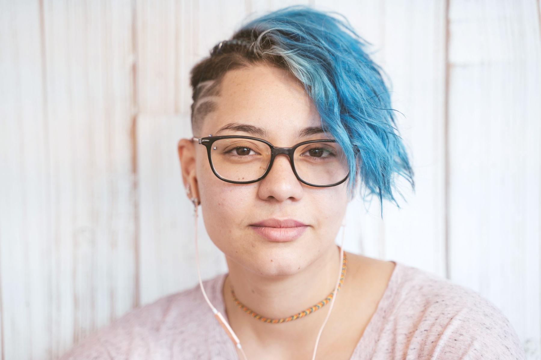 Close-up of a woman with short, blue hair with shaved sides, wearing glasses, earrings, a choker necklace, and pink shirt, sitting against a white wooden background.