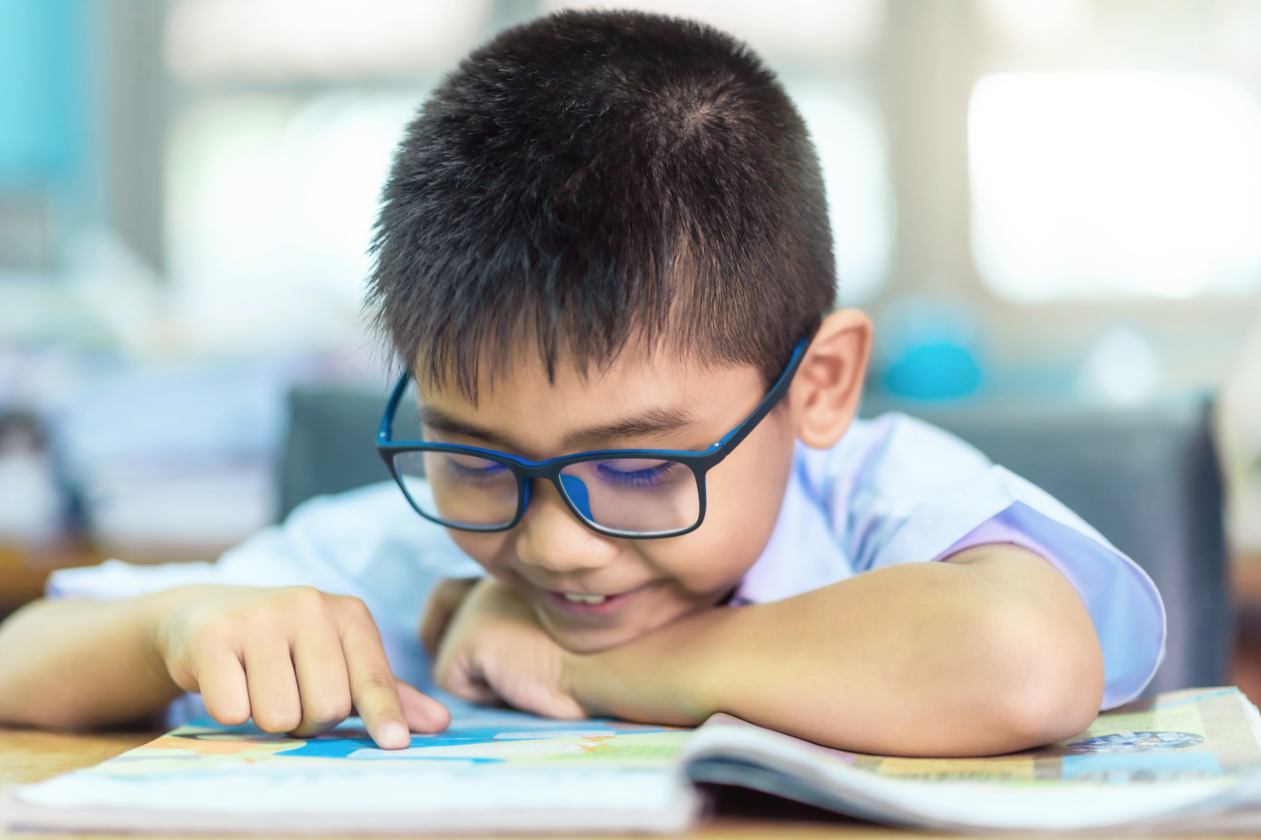 A young boy wearing glasses is reading a colorful book at a desk in a classroom.