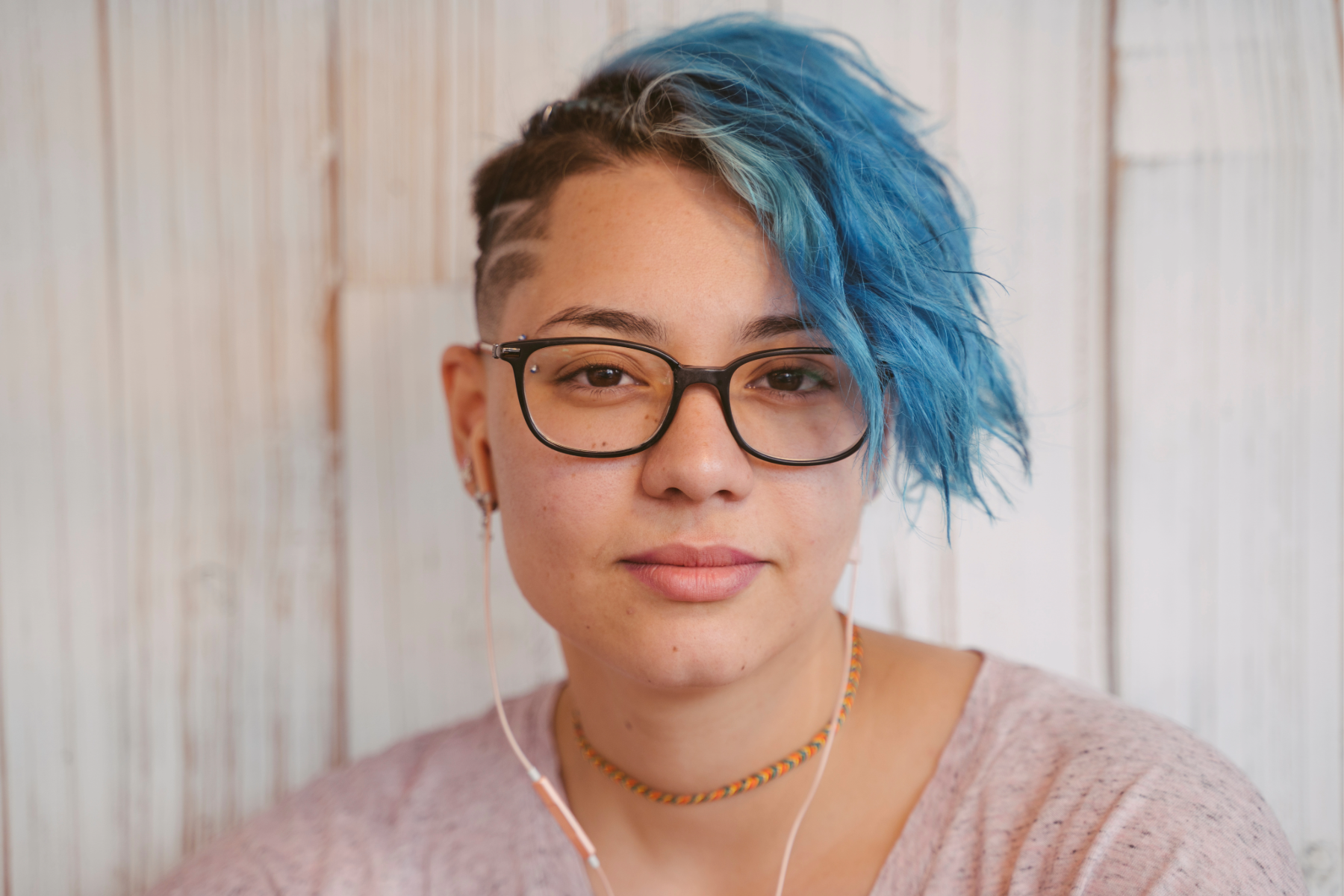 Close-up of a woman with short, blue hair with shaved sides, wearing glasses, earrings, a choker necklace, and pink shirt, sitting against a white wooden background.