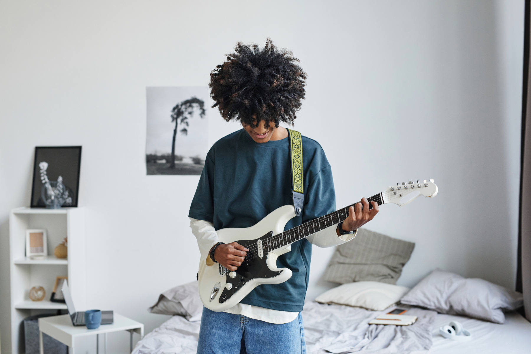 Young person with curly hair playing a white electric guitar in a bedroom.
