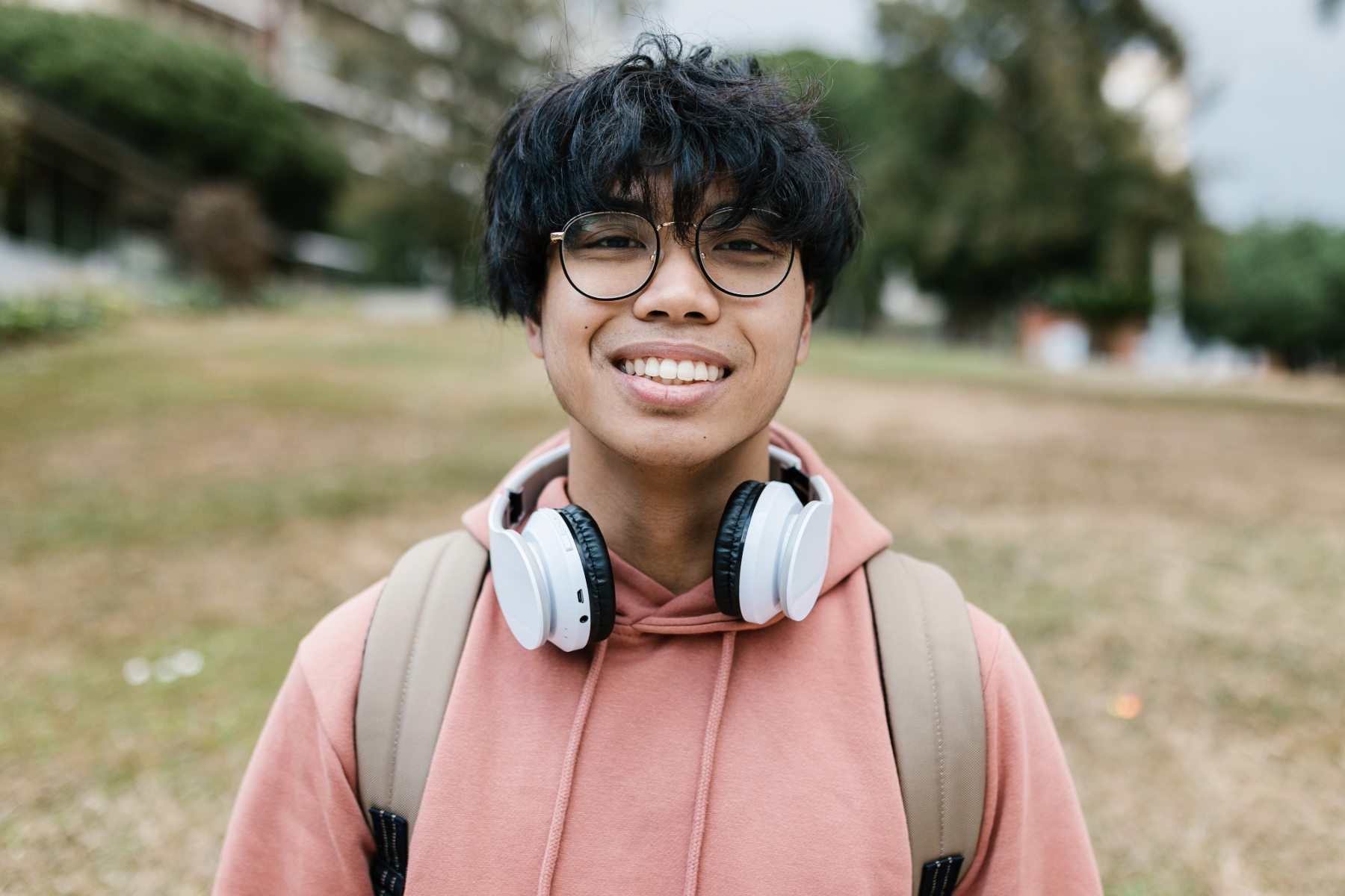 A young man outdoors smiling, wearing glasses, a pink hoodie, and carrying a backpack with white headphones around his neck.