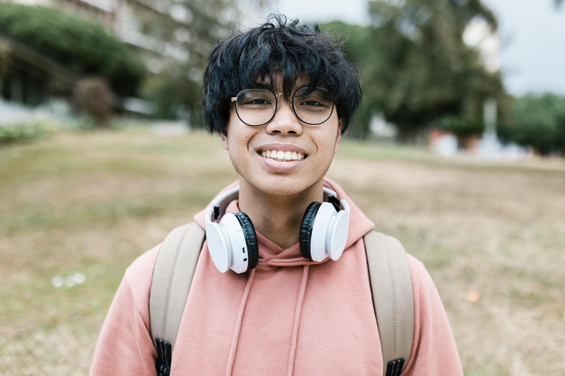 A young man outdoors smiling, wearing glasses, a pink hoodie, and carrying a backpack with white headphones around his neck.
