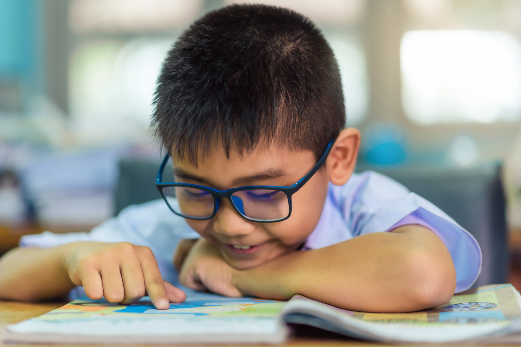 A young boy wearing glasses is reading a colorful book at a desk in a classroom.