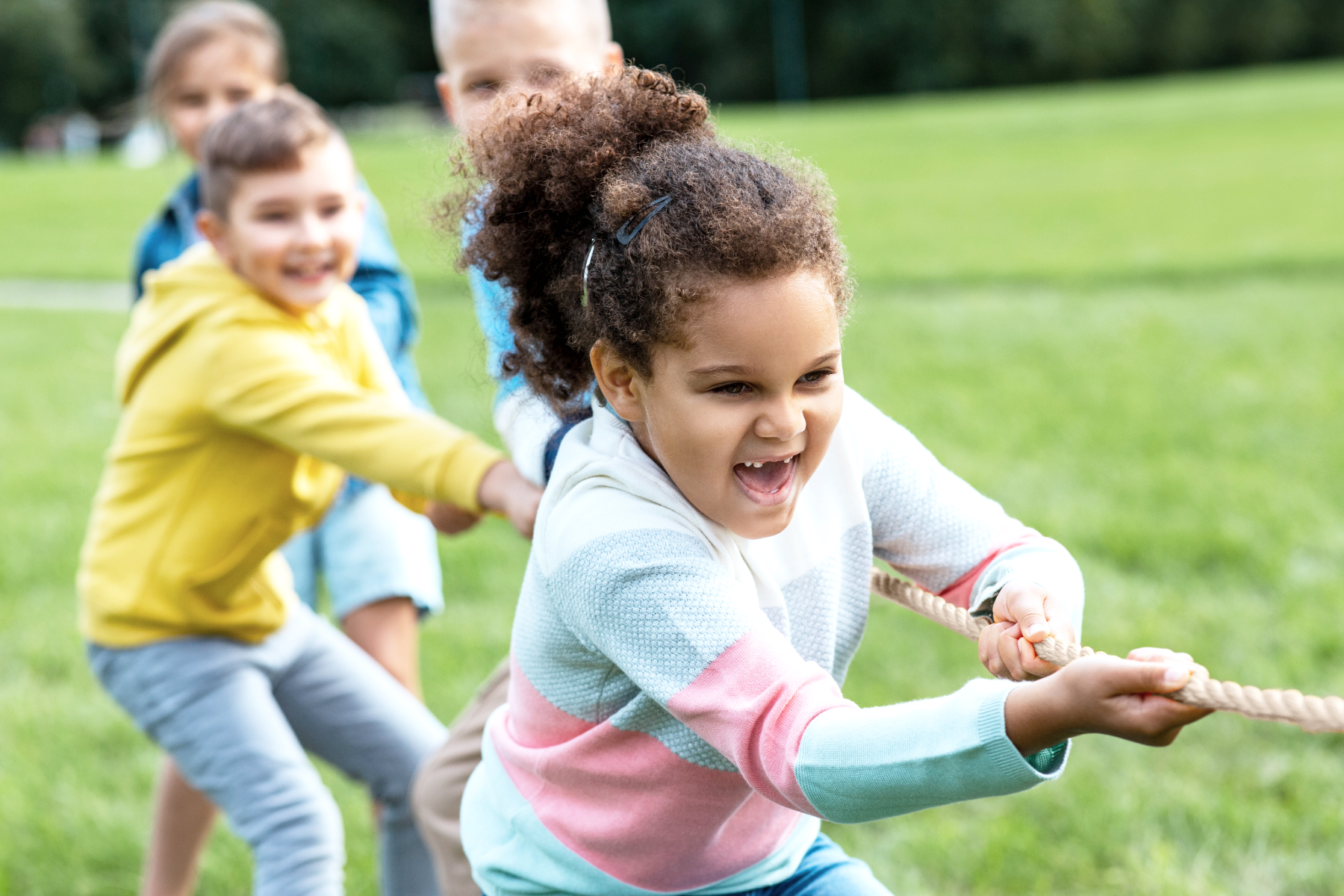 Four children playing tug of war outdoors on a grassy field, pulling the rope with expressions of effort and determination.