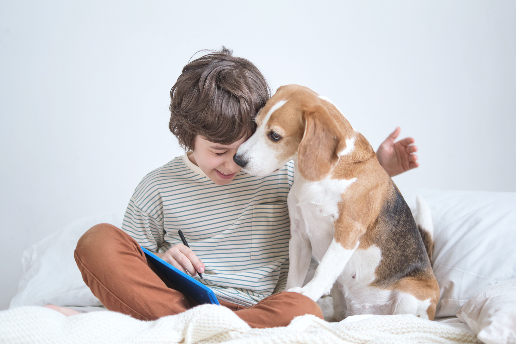 A young boy with brown hair, smiling, sitting on a bed with a beagle dog. The boy is writing or drawing on a notebook, and the dog is sniffing or gently touching his face.