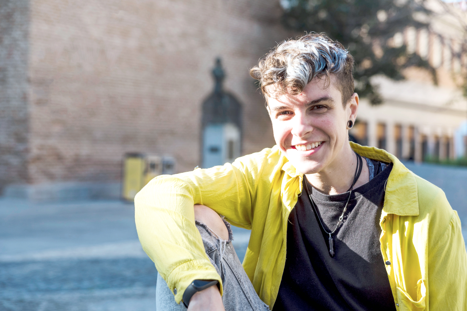 Young person with short curly hair, smiling, sitting outdoors in front of a brick wall, wearing a yellow jacket, black T-shirt, and accessories.