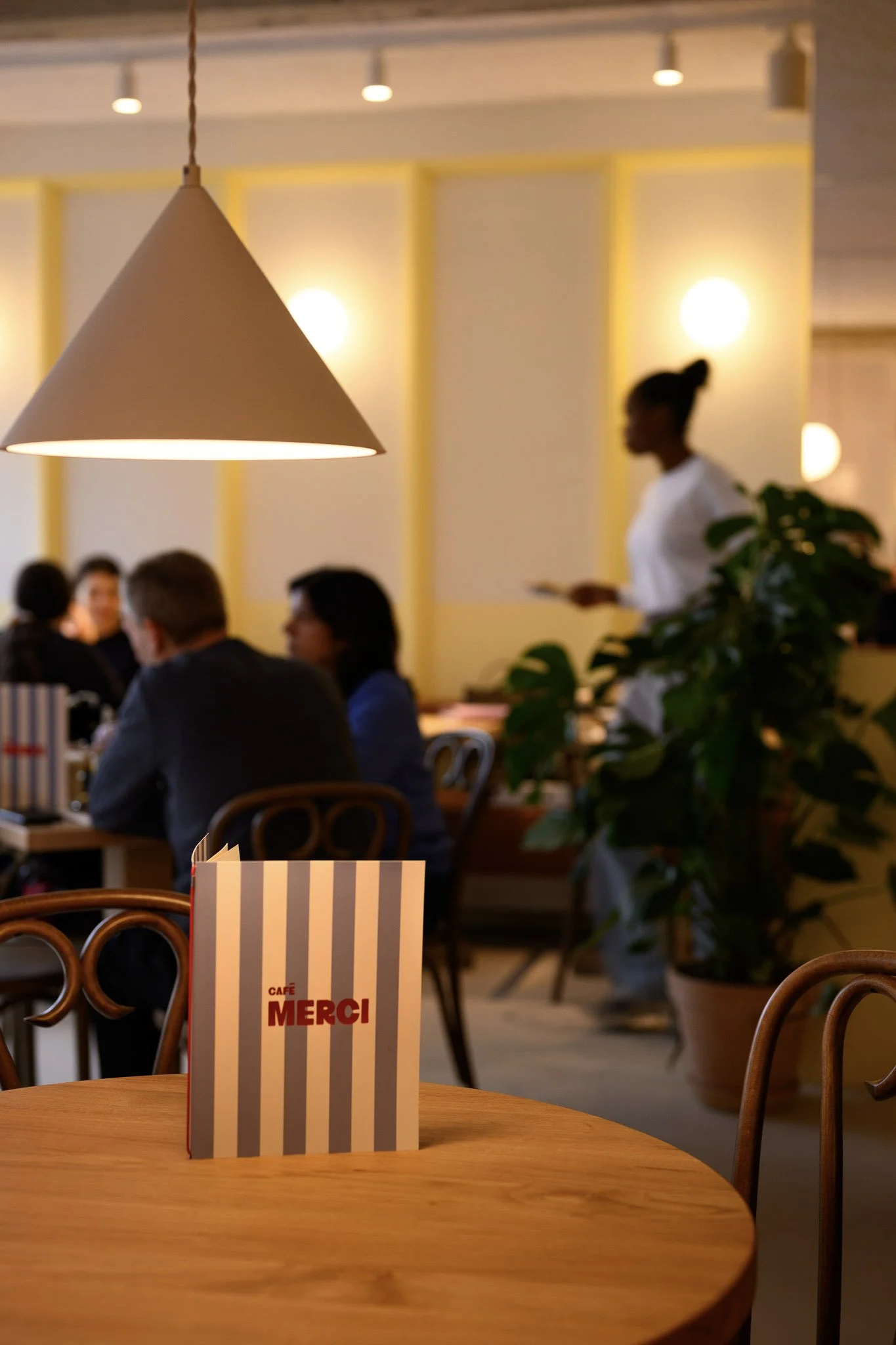 Een interieur van een restaurant met een houten tafel, een menu met de tekst 'Café Merci', een grote hanglamp en een groep mensen aan een tafel op de achtergrond, terwijl een serveerster voorbij loopt.