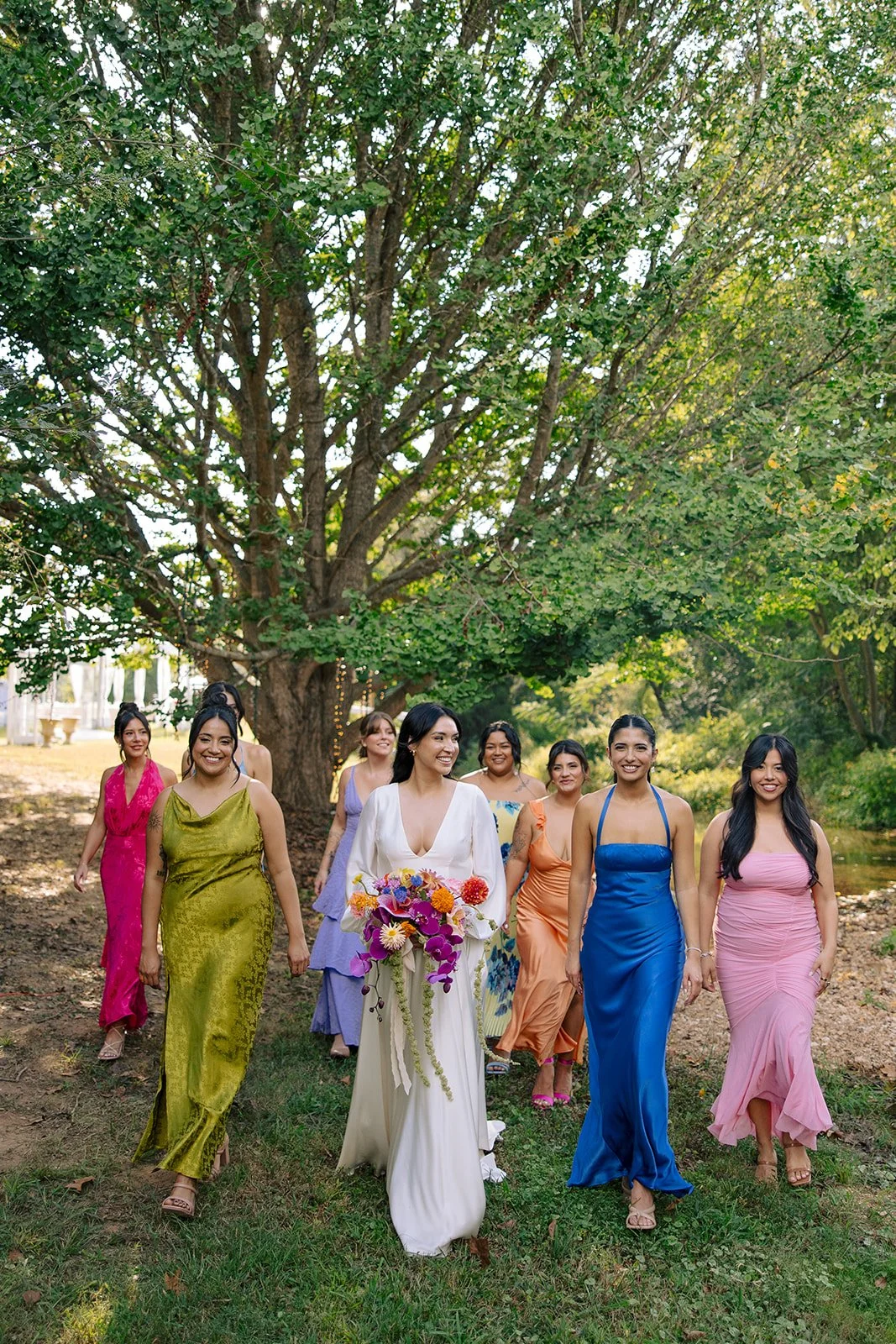 A group of women, including a bride in a white dress holding a bouquet, walking outdoors under a large tree in a lush green setting, possibly at a wedding or celebration.