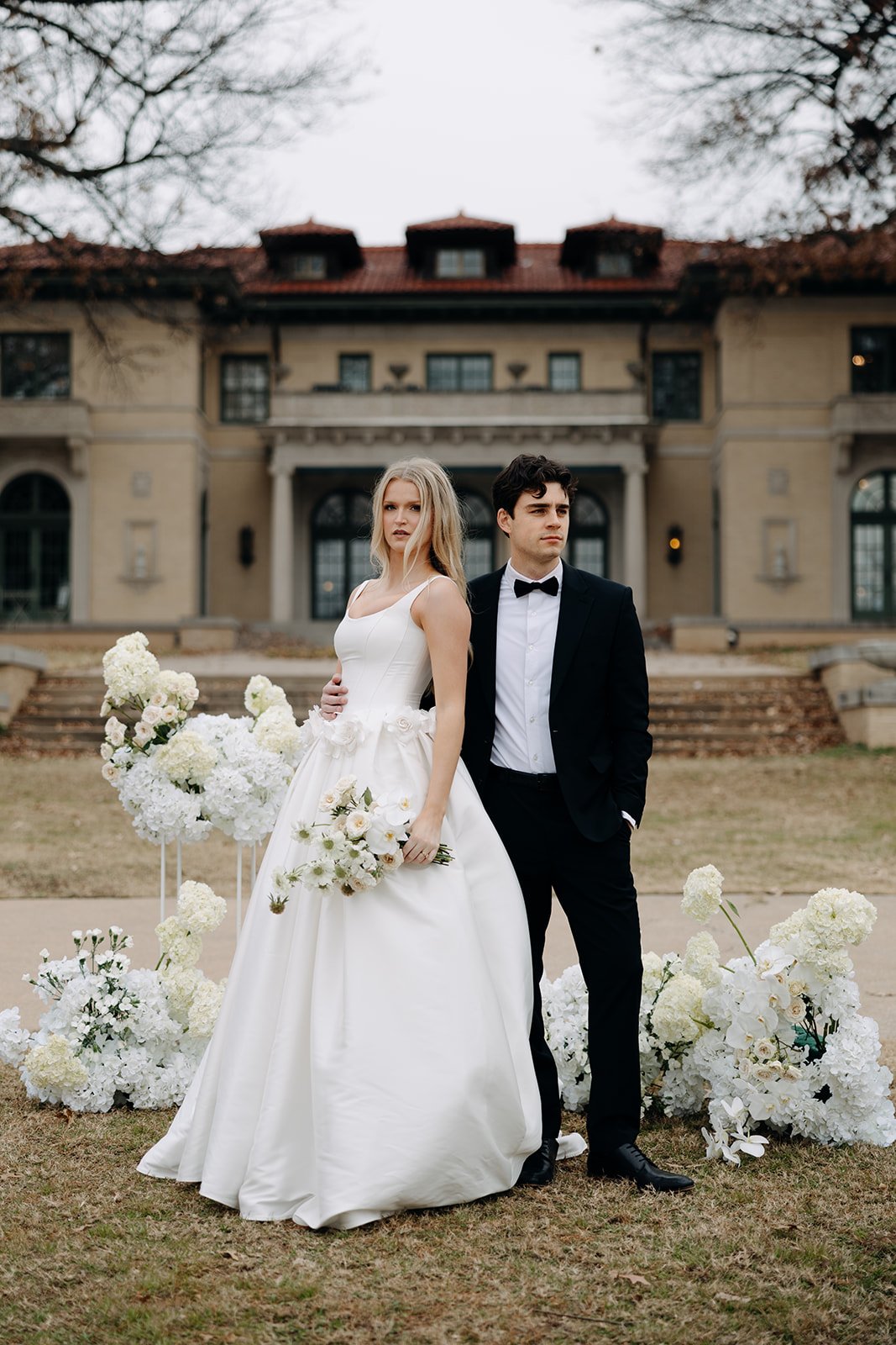 A couple dressed in wedding attire standing outdoors in front of a large house, surrounded by floral arrangements.