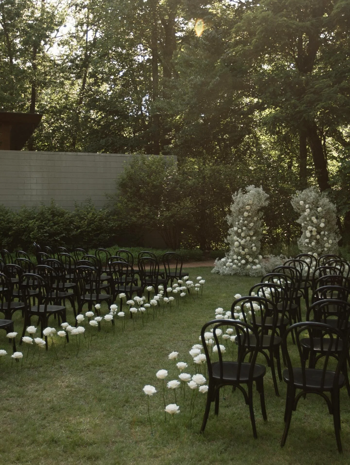 Outdoor wedding ceremony setup with black chairs arranged on grass, white floral decorations on the ground, and two large flower arches in the background surrounded by trees.
