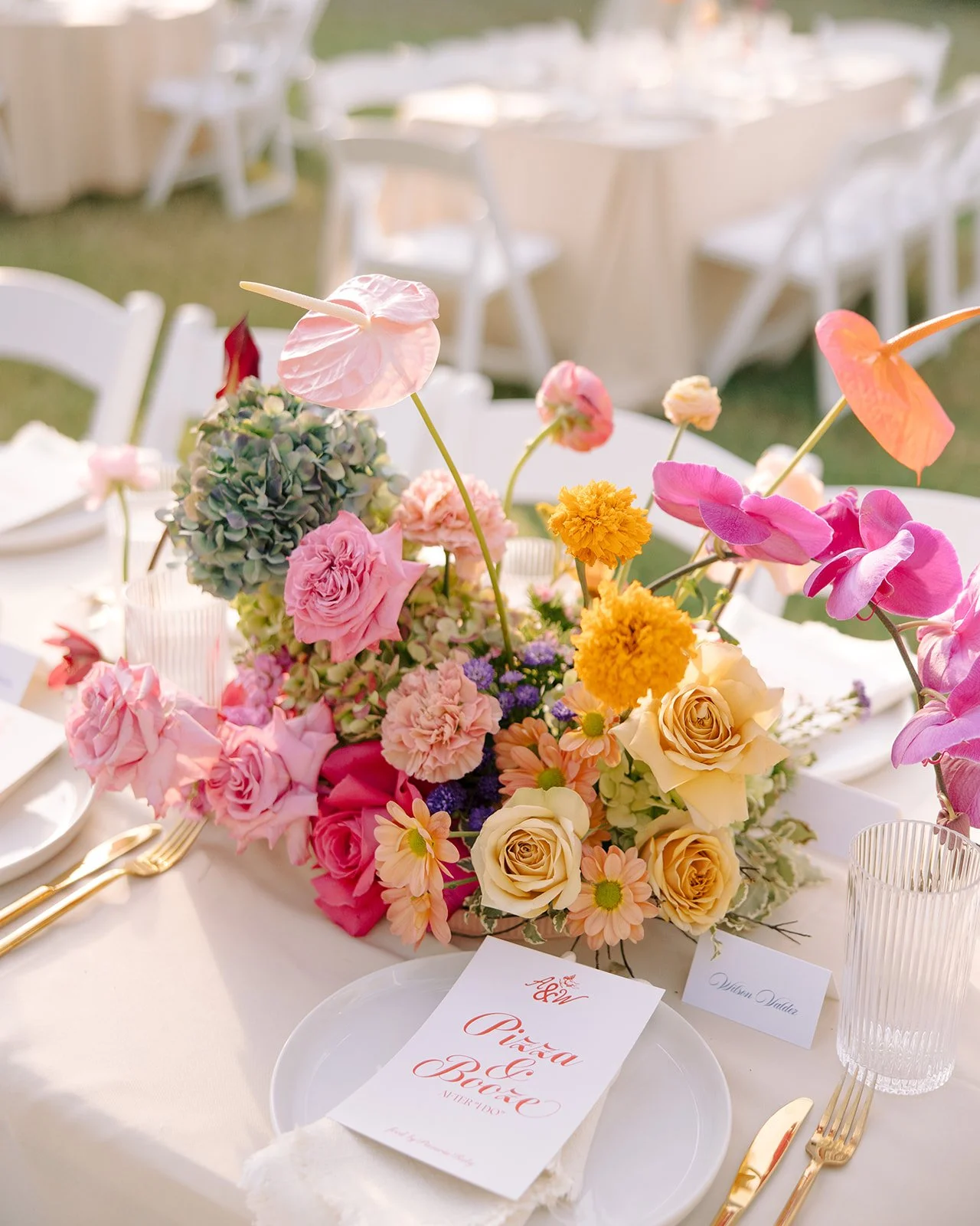 Colorful floral centerpiece on a white table at a wedding reception with gold cutlery, white plates, and pink anthuriums.