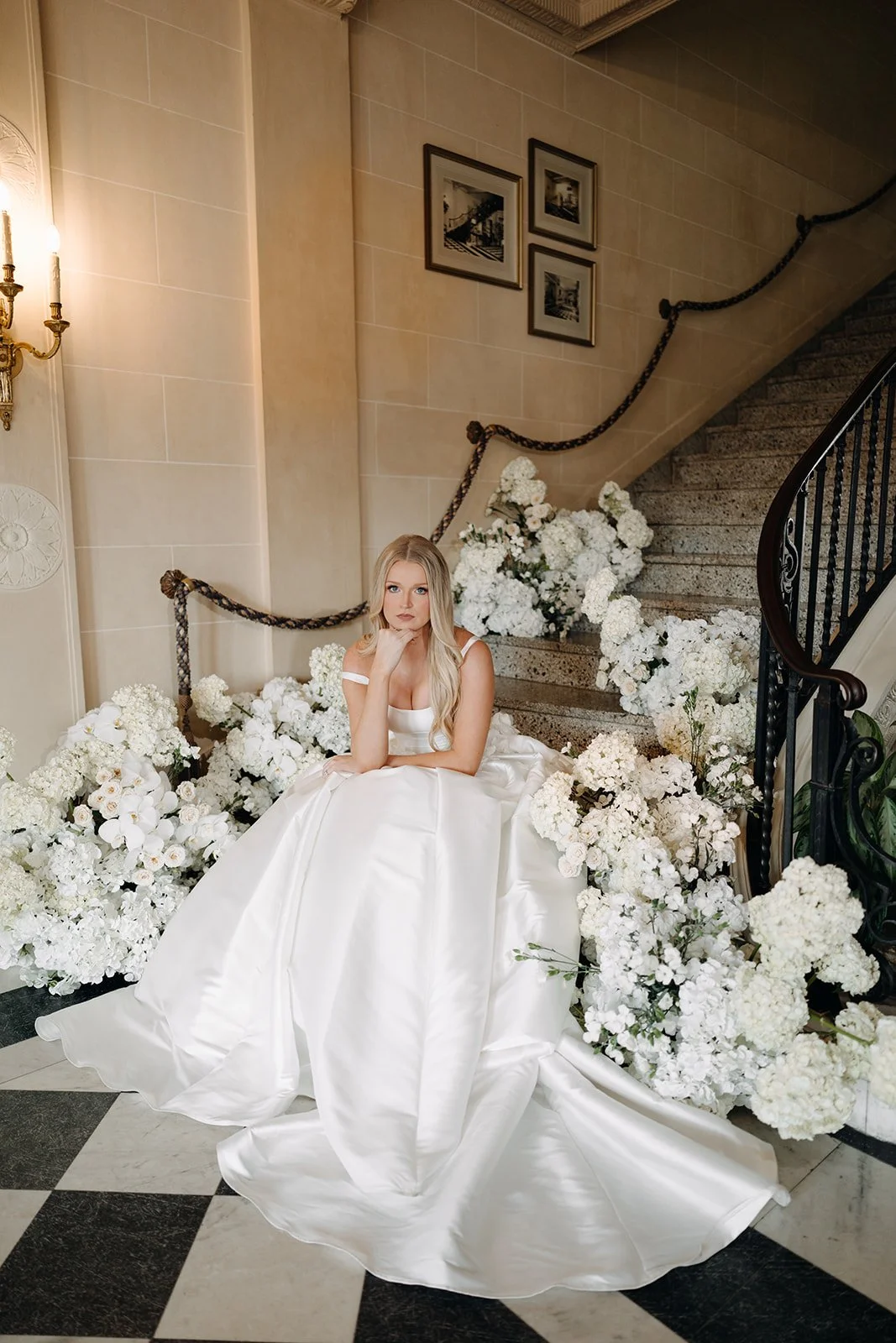 A woman in a white wedding dress sits on the stairs surrounded by white flowers with a marble staircase in the background.