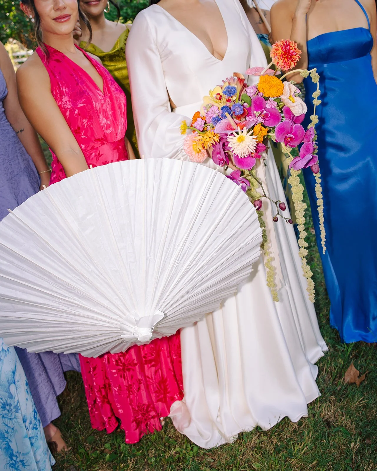 Group of women dressed in colorful dresses, one holding a bouquet of vibrant flowers and parasol, at an outdoor event