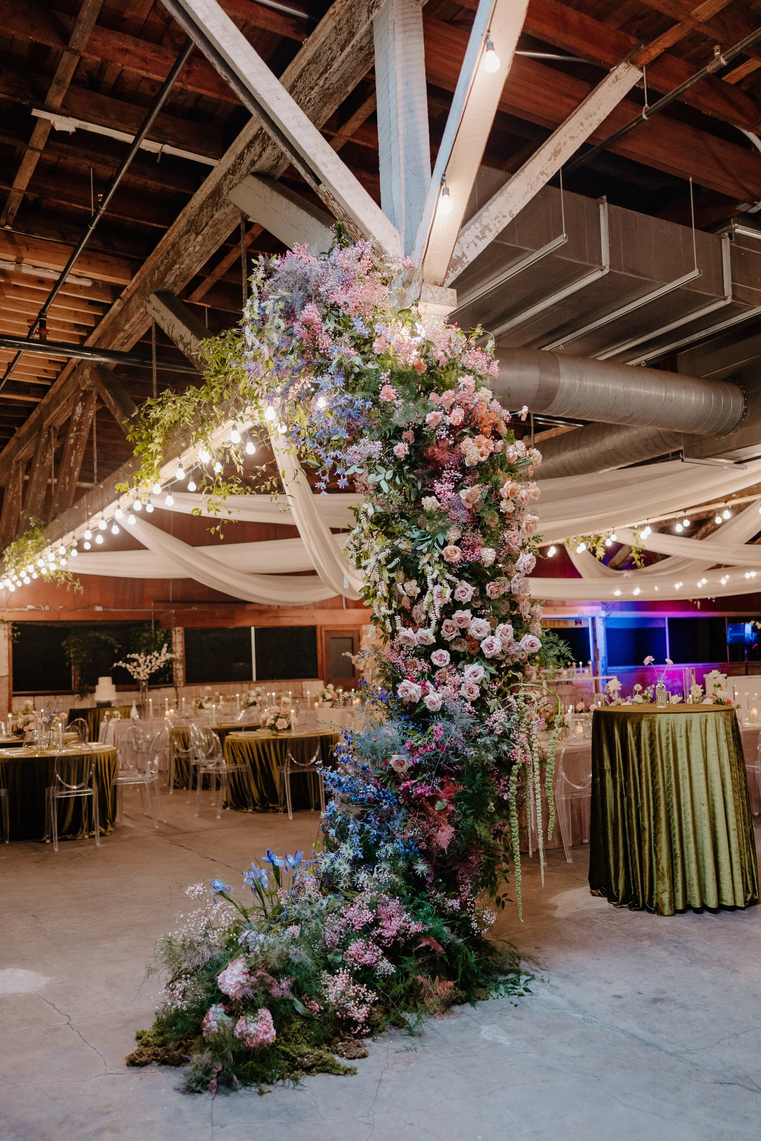 A tall floral wedding arch decorated with pink and purple flowers, greenery, and draped fabric, inside a rustic event venue with string lights, wooden beams, and tables with elegant decor.