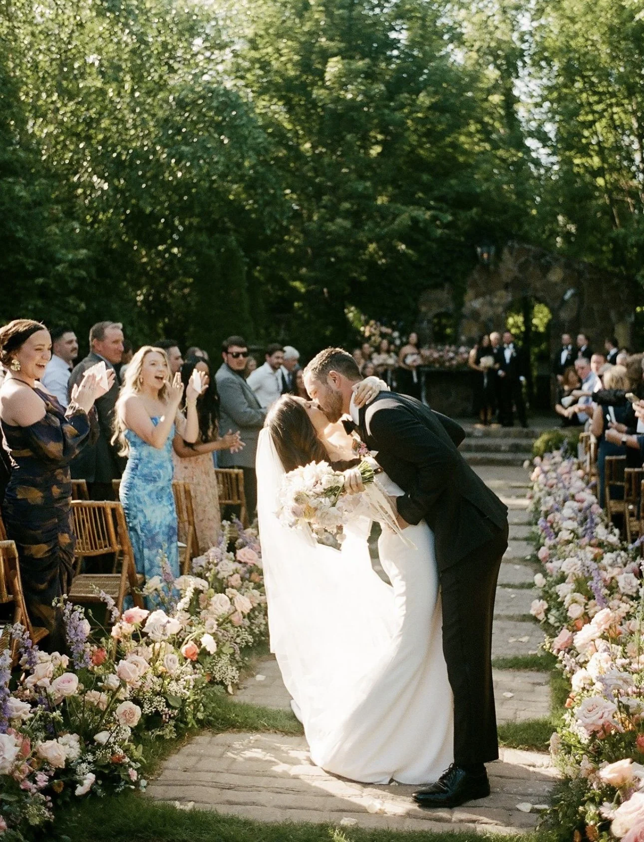 Bride and groom sharing a kiss during outdoor wedding ceremony, surrounded by guests and decorated with flowers.