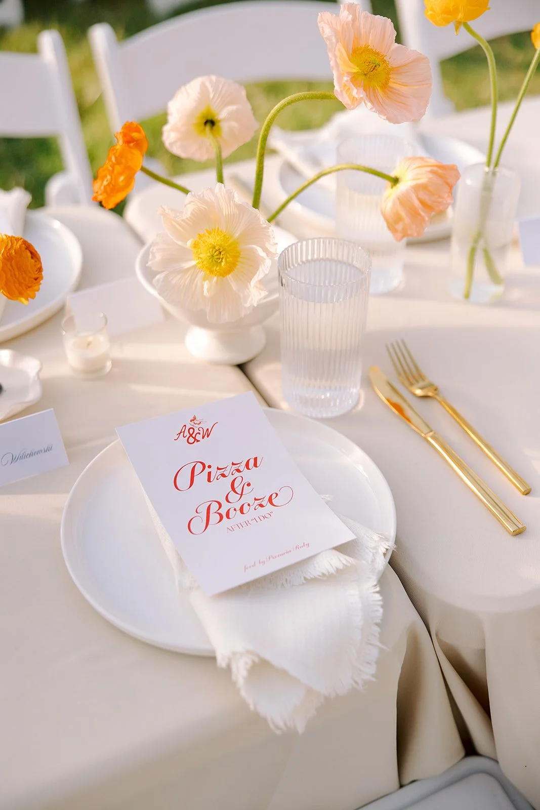 Table setting with white plates, gold cutlery, clear glasses, a menu card, and a floral centerpiece with pastel-colored poppies in a white vase at an outdoor event.