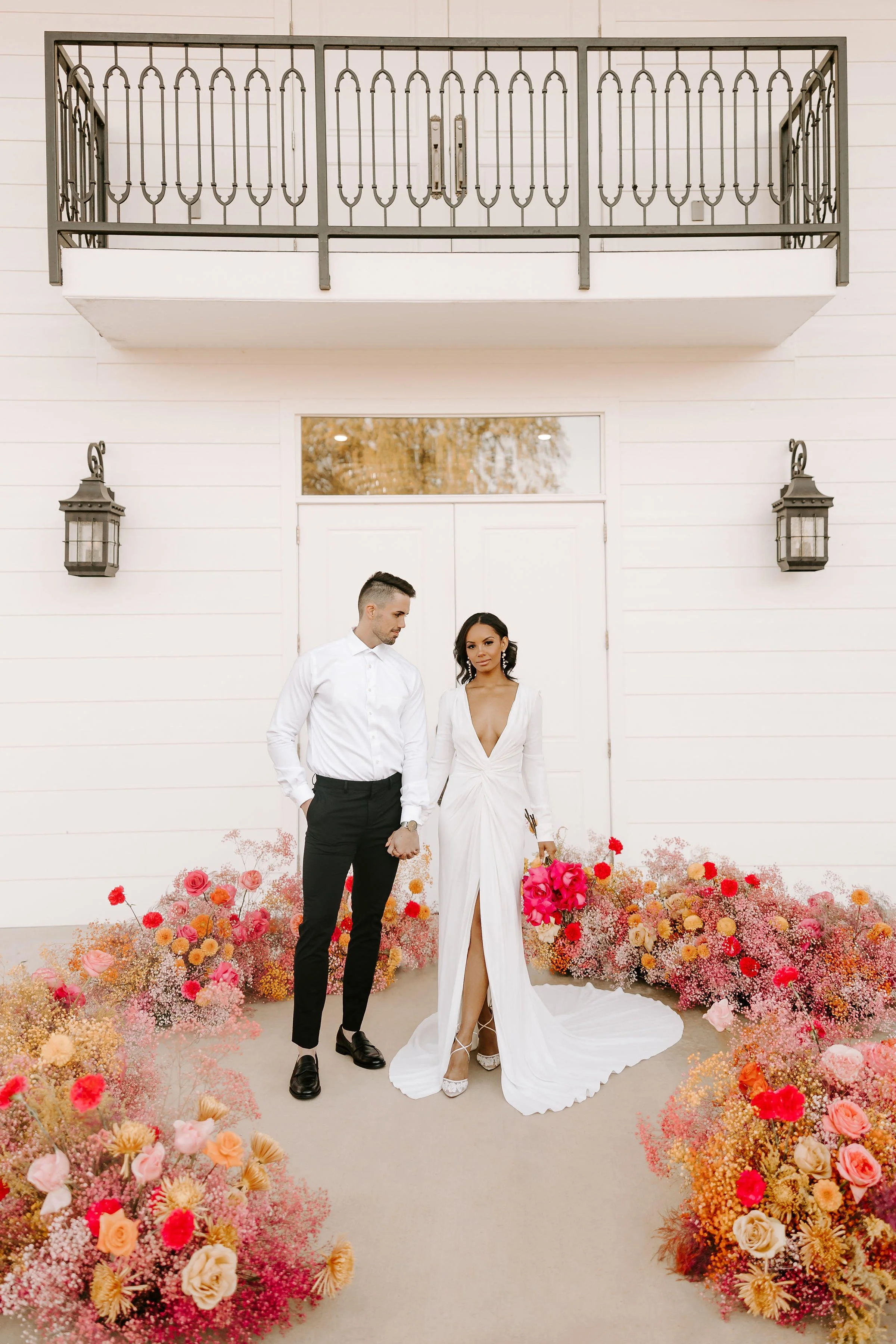 A stylish couple dressed in wedding attire, holding hands inside a vibrant flower arch, standing in front of a white building with a balcony and lanterns.