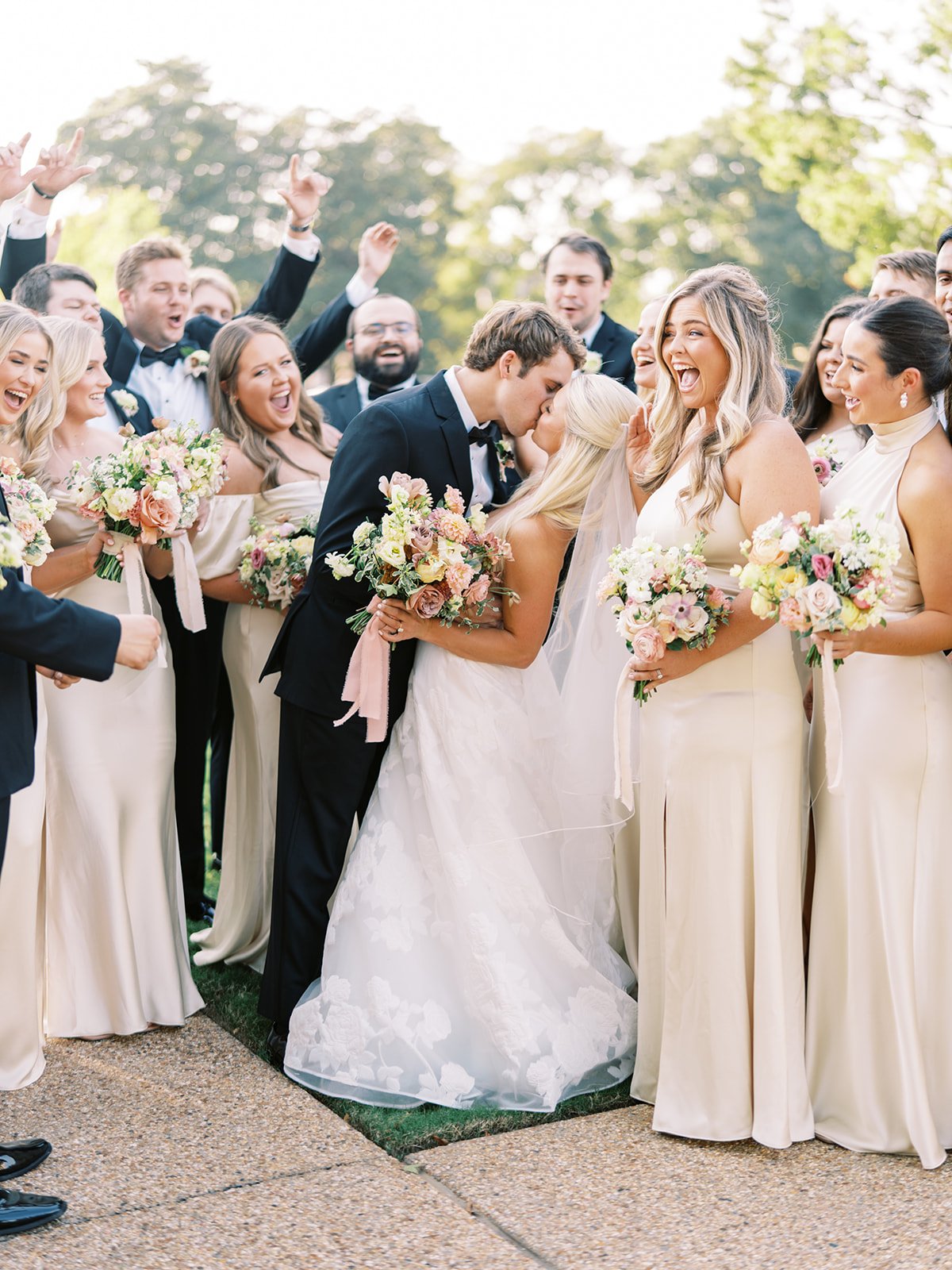 A wedding celebration with a bride and groom kissing, surrounded by bridesmaids and groomsmen outdoors, all smiling and holding bouquets of flowers.
