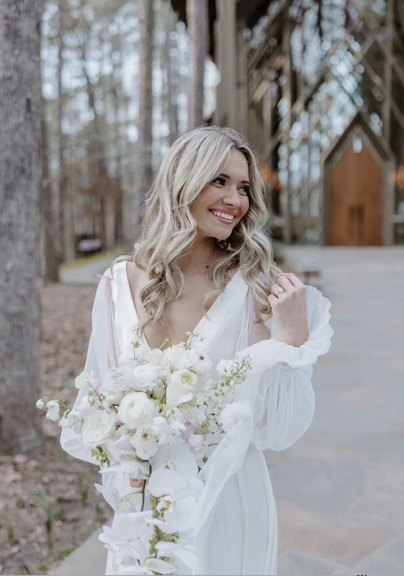 A smiling woman in a white dress holding a bouquet of white flowers, outdoors among trees with a wooden building in the background.