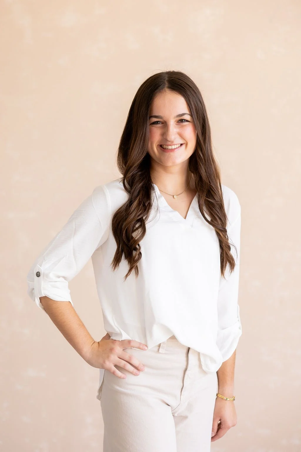 A young woman with long brown hair, wearing a white blouse with rolled-up sleeves and beige pants, smiling and standing with her hand on her hip against a plain beige background.