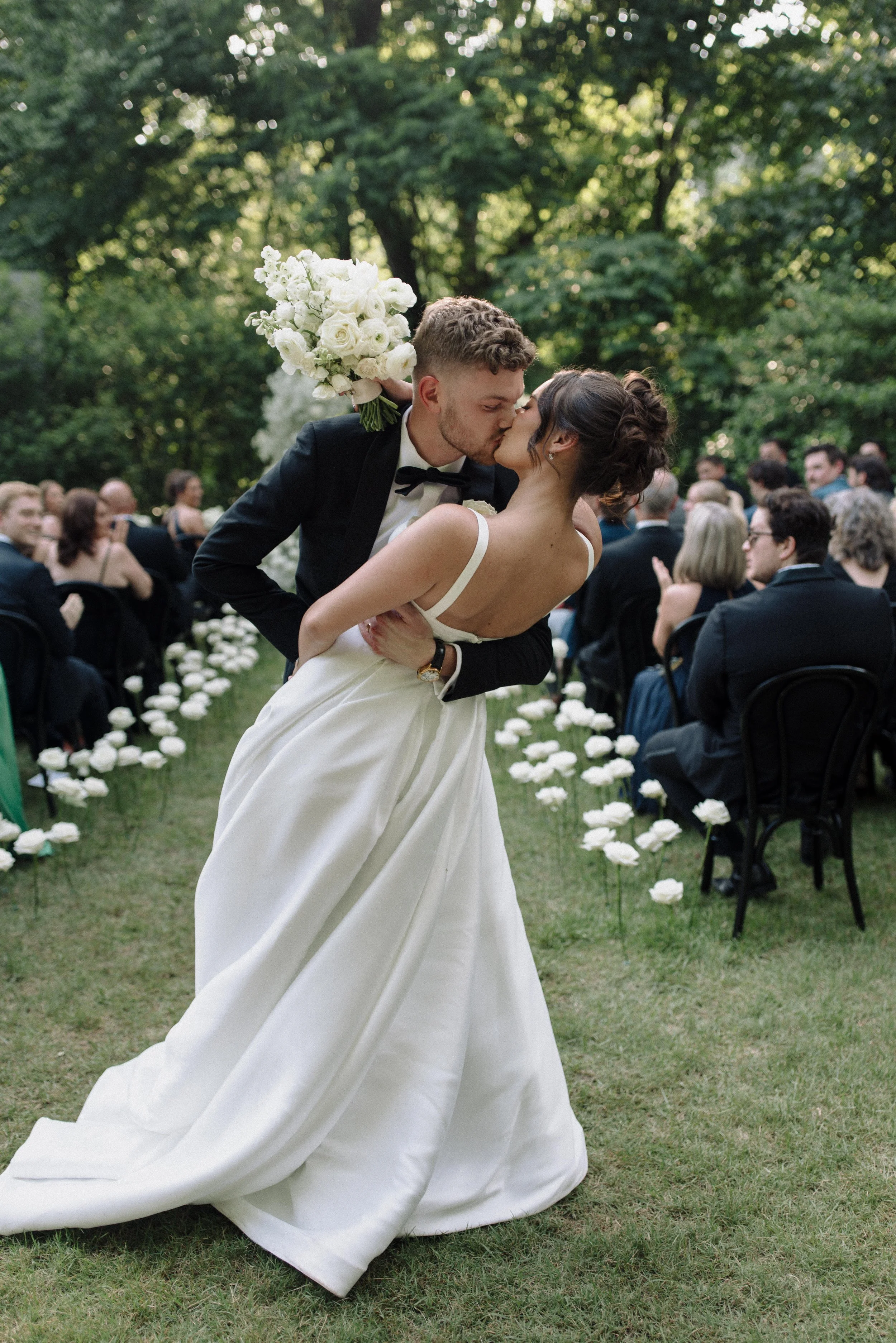 A bride and groom kissing during their outdoor wedding ceremony, surrounded by guests seated on black chairs, with white flower decorations on the grass, and lush green trees in the background.