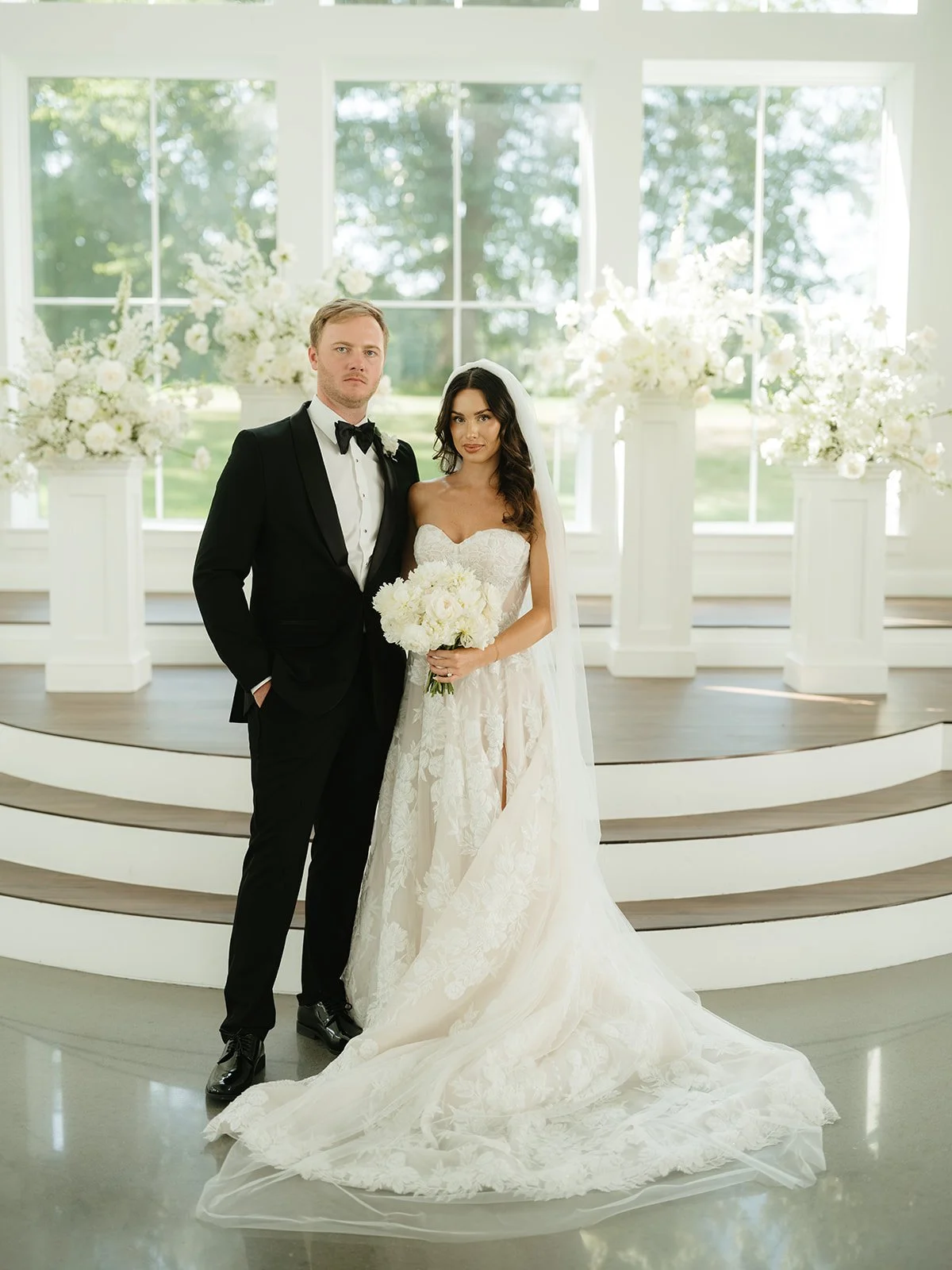 A bride and groom standing together on a wedding stage with large floral arrangements in the background, inside a bright and airy venue.