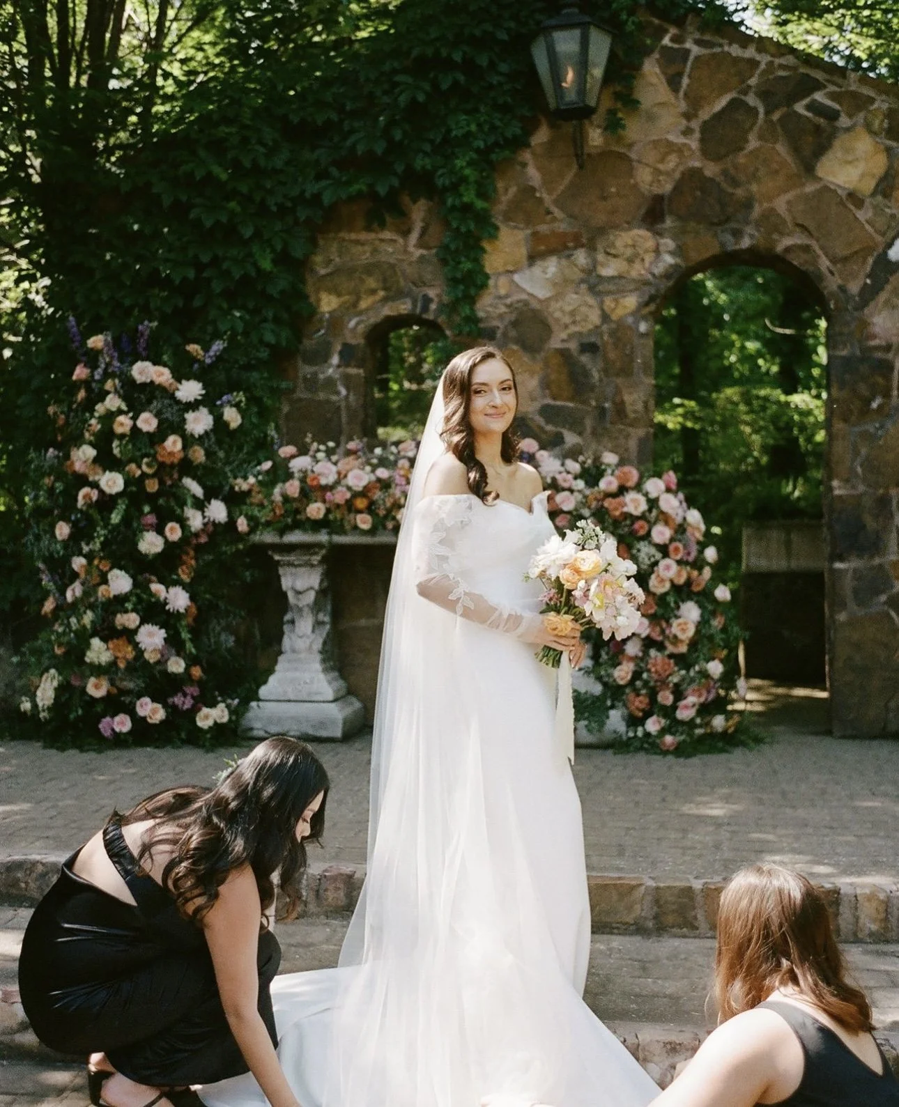 A bride in a white wedding dress holding a bouquet of flowers, smiling, with two women adjusting the train of her dress. She is standing outdoors near a stone structure with greenery and pink flowers in the background.