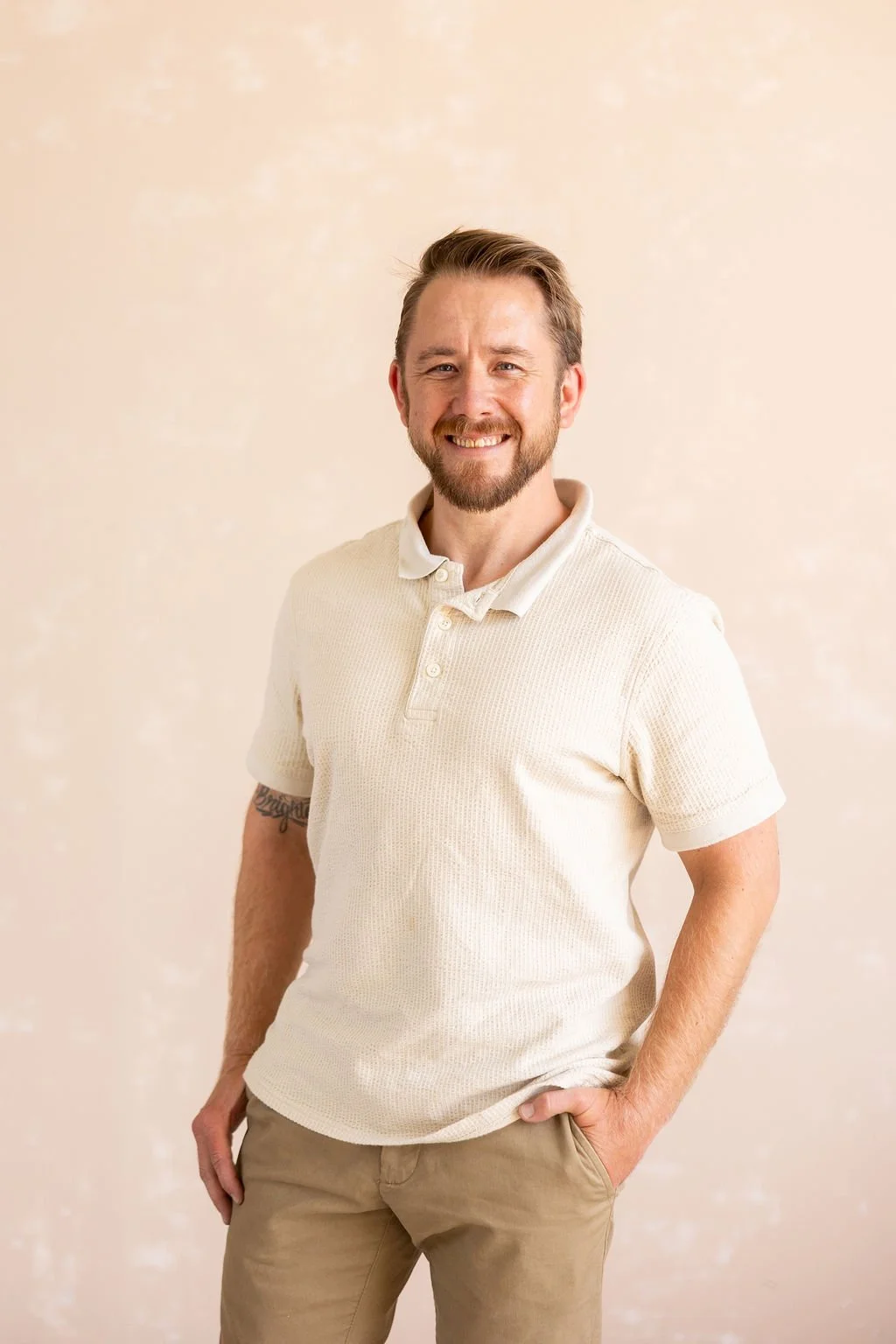 A smiling man with a beard wearing a cream-colored short-sleeve polo shirt and khaki pants, standing against a light beige background.