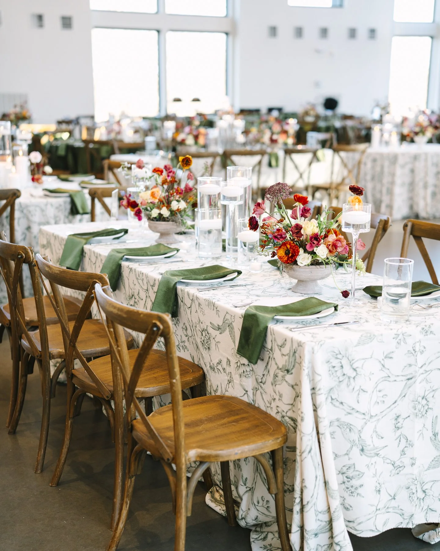 A decorated event table with floral centerpieces, glass candle holders, green napkins, and tableware inside a well-lit venue with large windows.