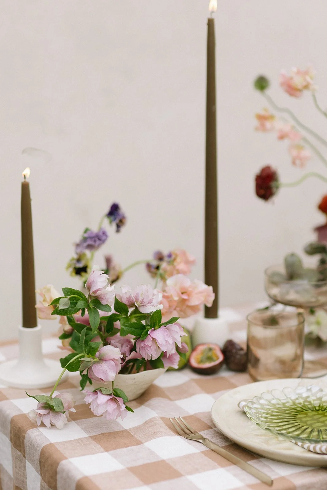 Table setting with pink and purple flowers, two lit candles, a sliced passion fruit, a green patterned plate, and a fork on a checkered tablecloth.