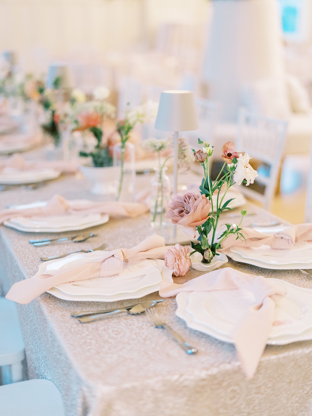 Elegant table setting with white plates, pink cloth napkins, silverware, and floral centerpieces with pink and white flowers on a lace tablecloth.