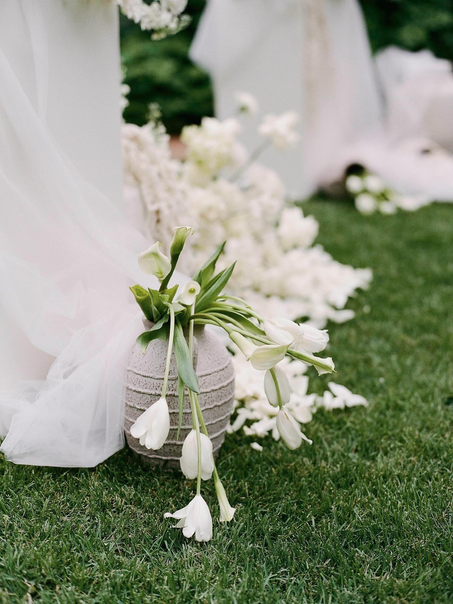 White flower arrangement with calla lilies and other white flowers in a textured vase on grass, with white drapery and additional flowers in the background.