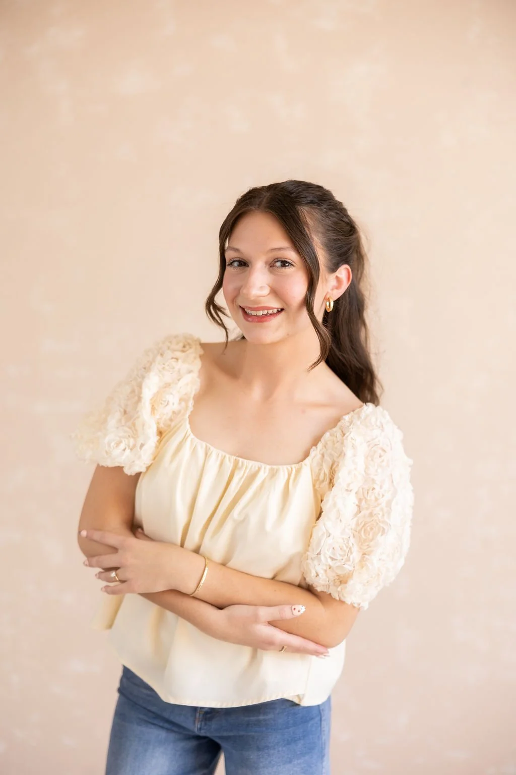 A young woman with brown hair styled in loose waves, wearing a cream-colored top with floral puff sleeves, blue jeans, and gold jewelry, smiling with her arms crossed against a beige background.