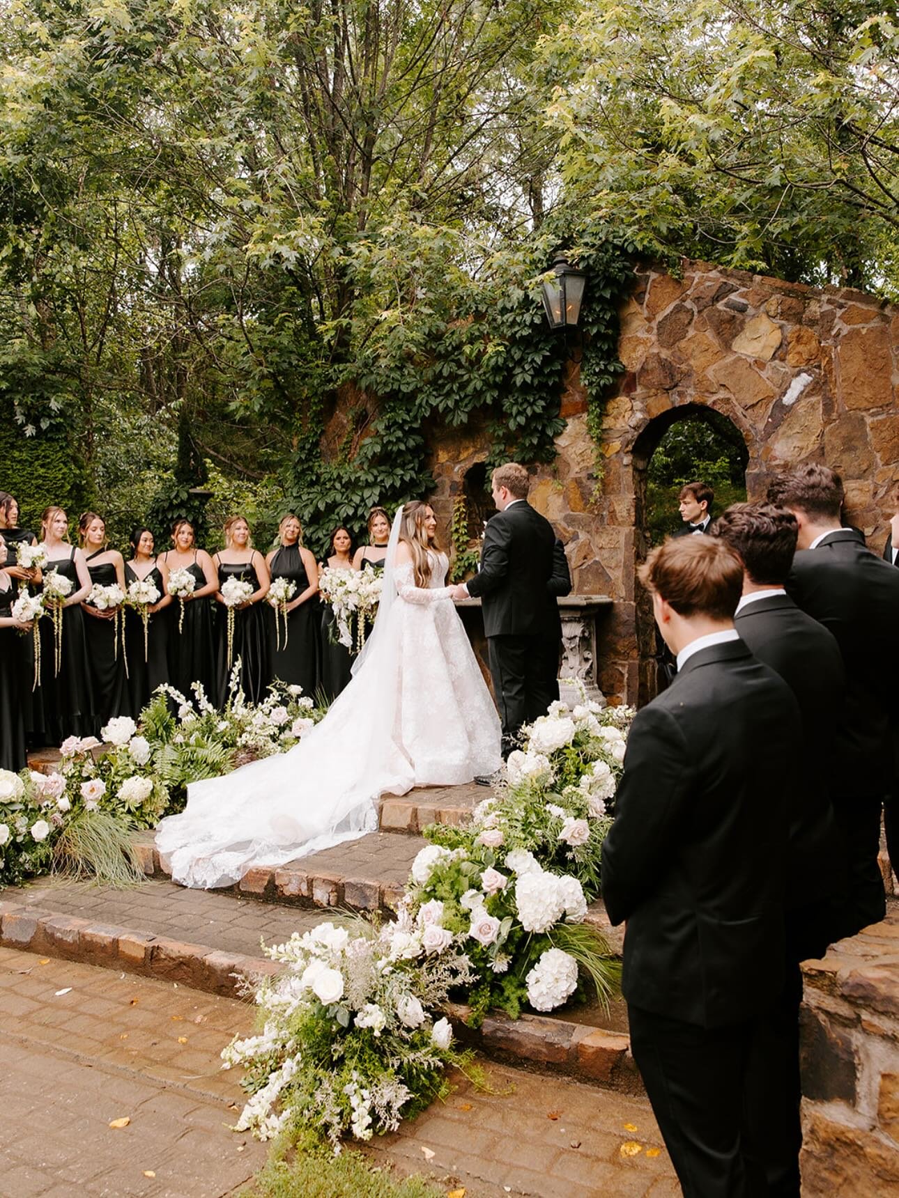 A wedding ceremony with a bride and groom exchanging vows outdoors surrounded by bridesmaids and groomsmen, with flowers and greenery.