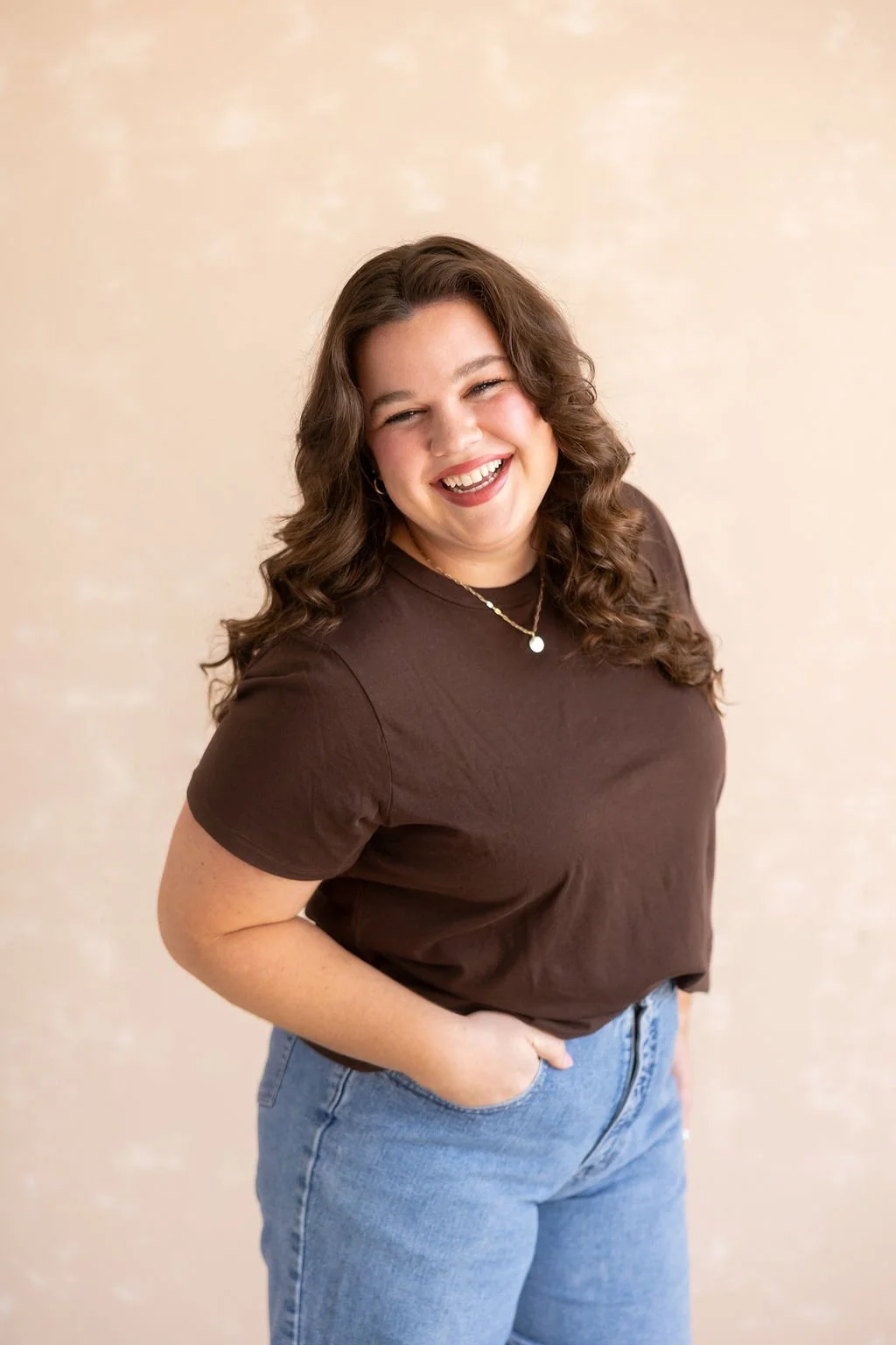 A woman with brown, curly hair smiling and wearing a brown t-shirt and blue jeans, standing against a neutral background.