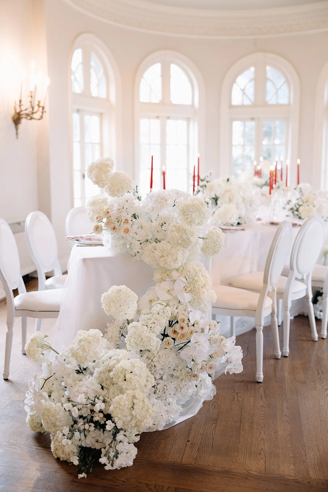 Elegant wedding table centerpiece with white flowers, including hydrangeas and roses, decorated with tall red candles in a brightroom with large arched windows.